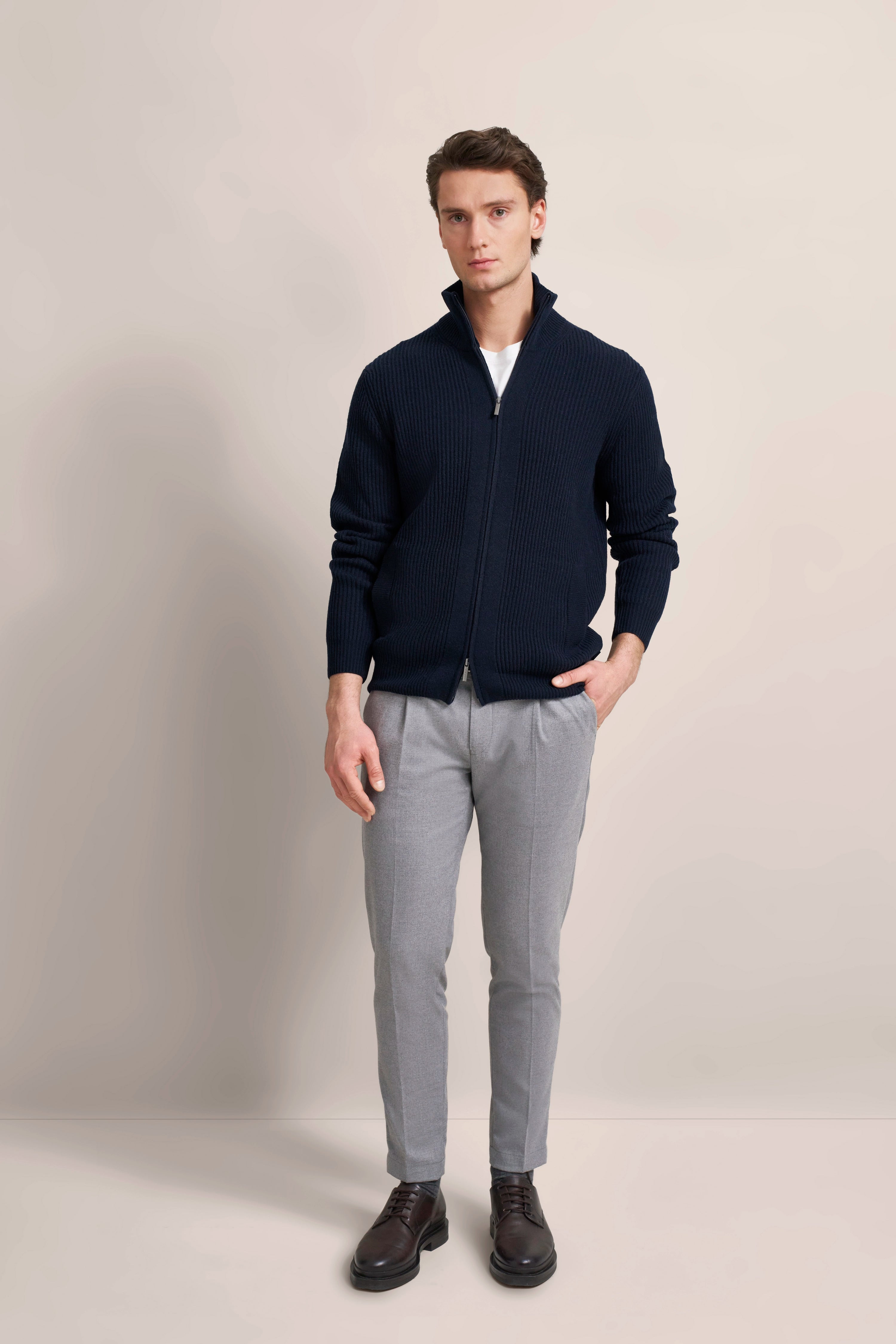 Model in bugatti-Outfit: Dunkelblauer Strickpullover, graue Hose, braune Schuhe. Entspanntes Posing.