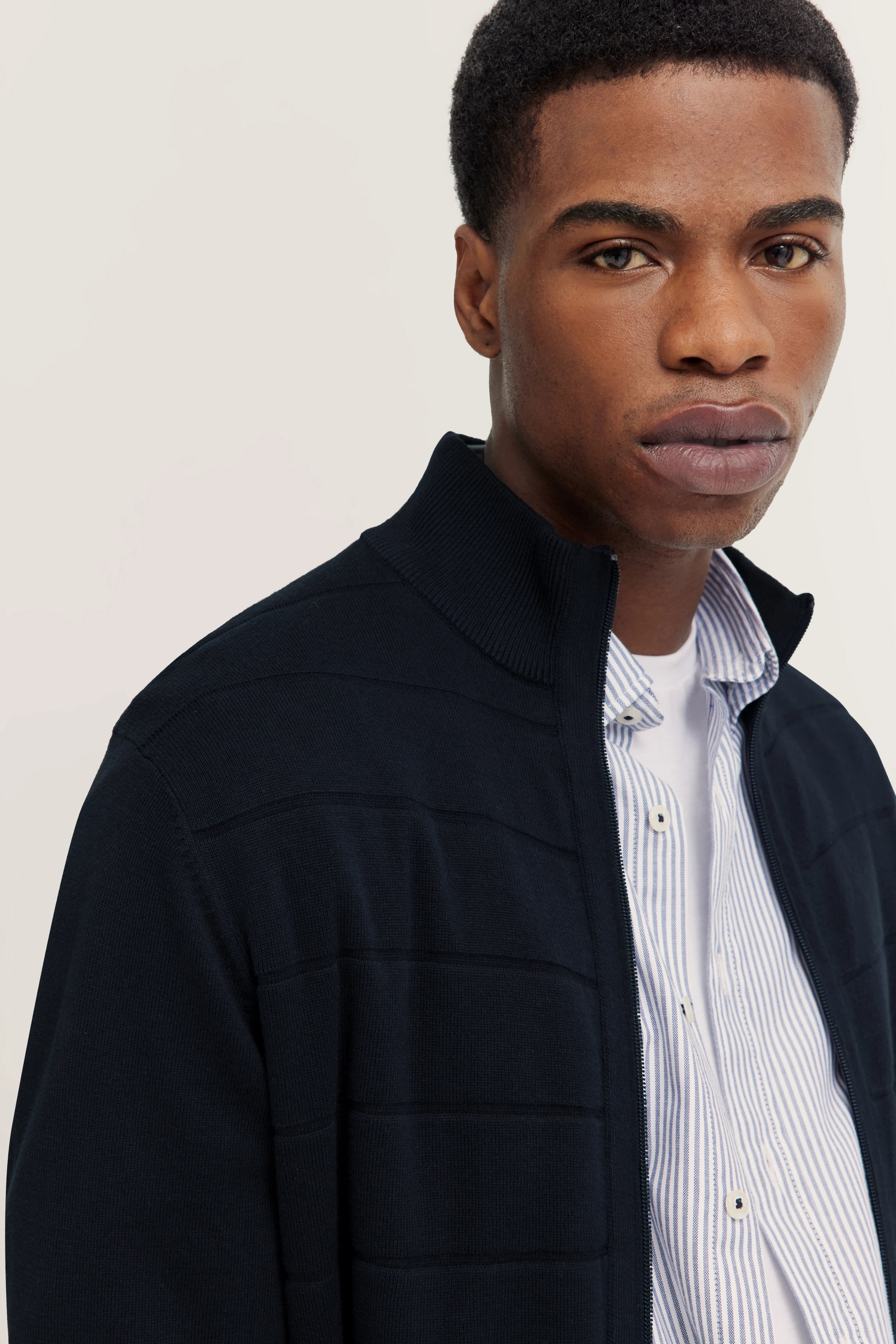 Model trägt bugatti Pullover in Dunkelblau mit Stehkragen, darunter ein gestreiftes Hemd.