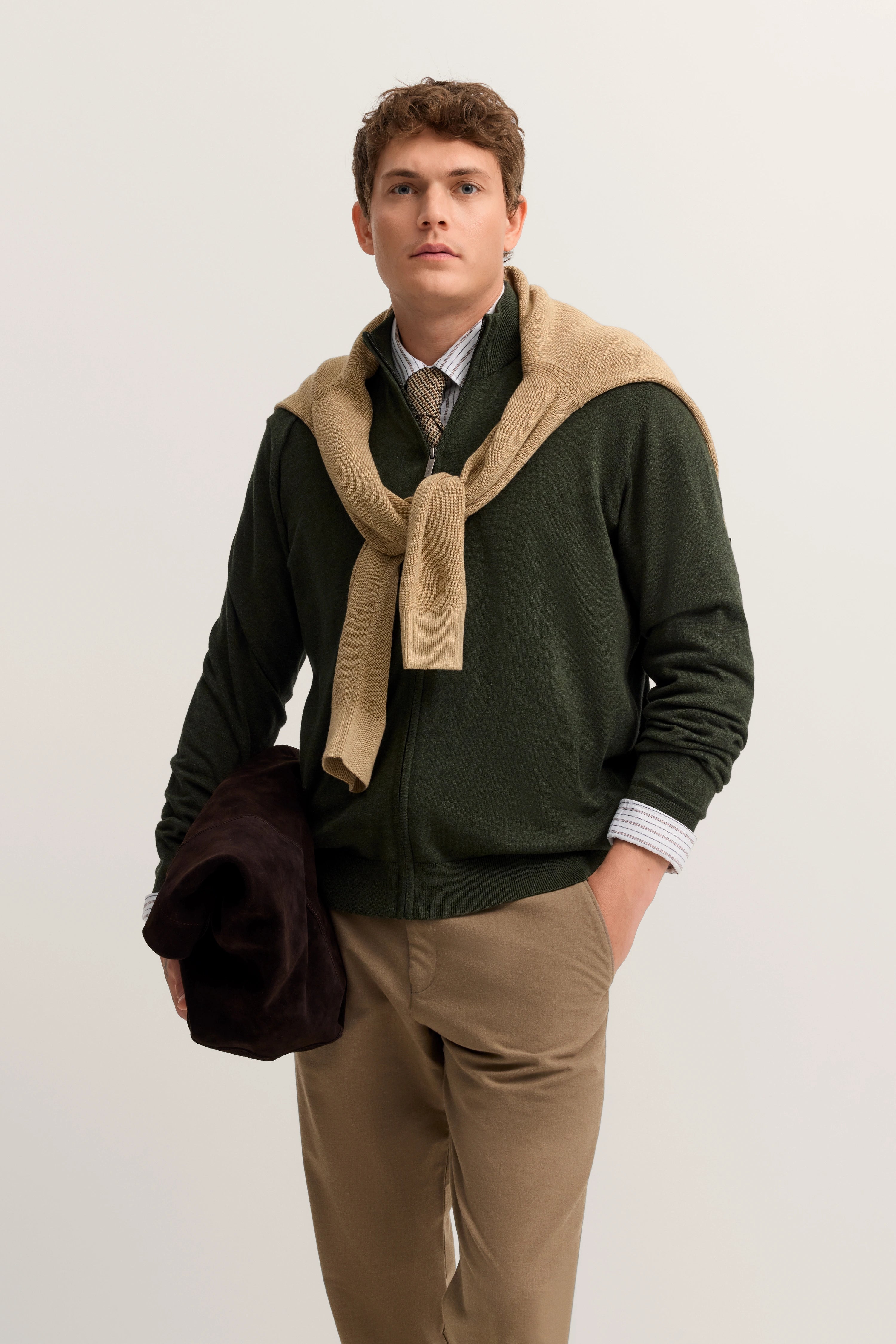 Model in olivgrüner Jacke, beige Hose, Hemd, Pullover über Schultern, lässige Pose.