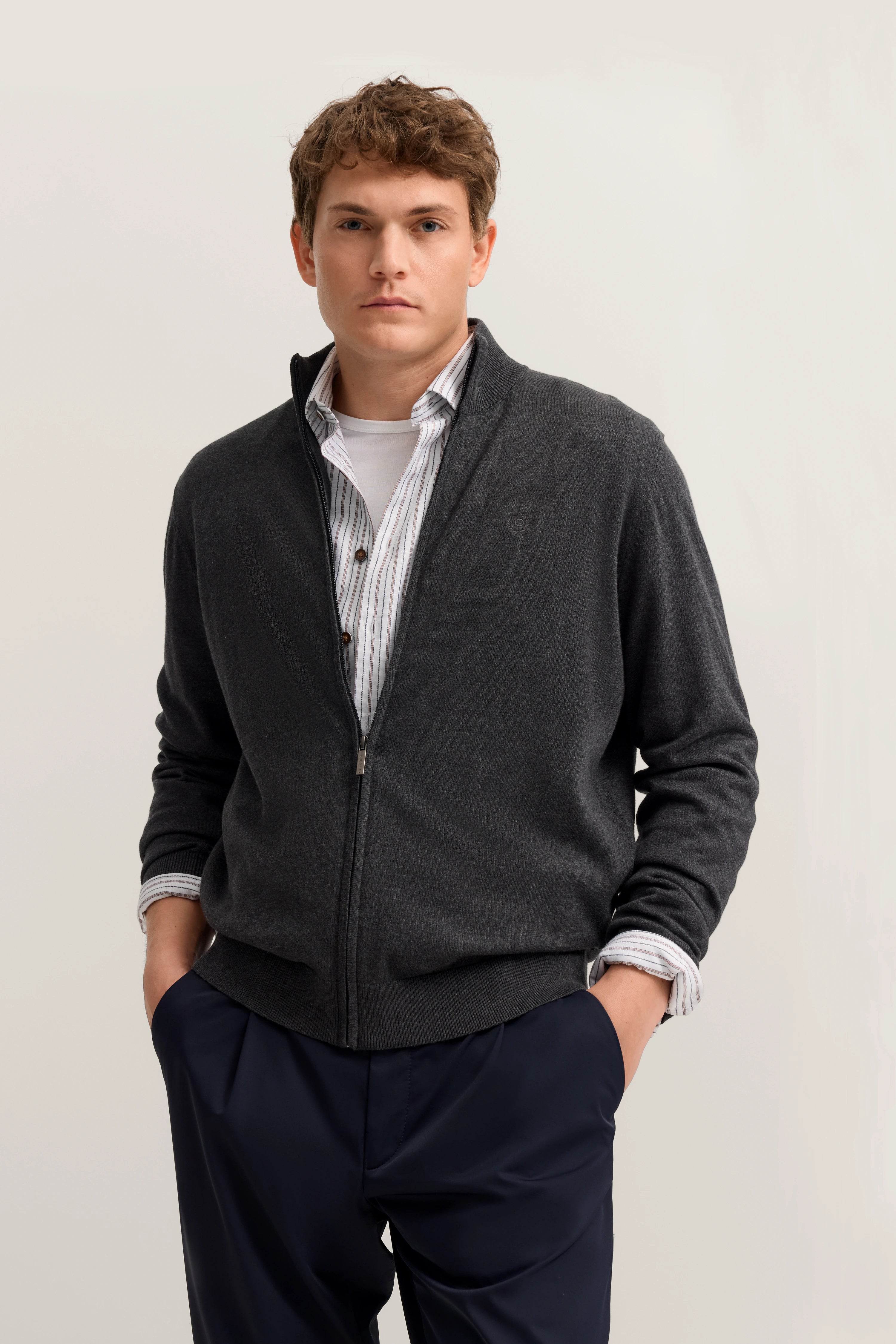 Grauer bugatti Cardigan über gestreiftem Hemd, dunkle Hose, Model mit entspanntem Posing.