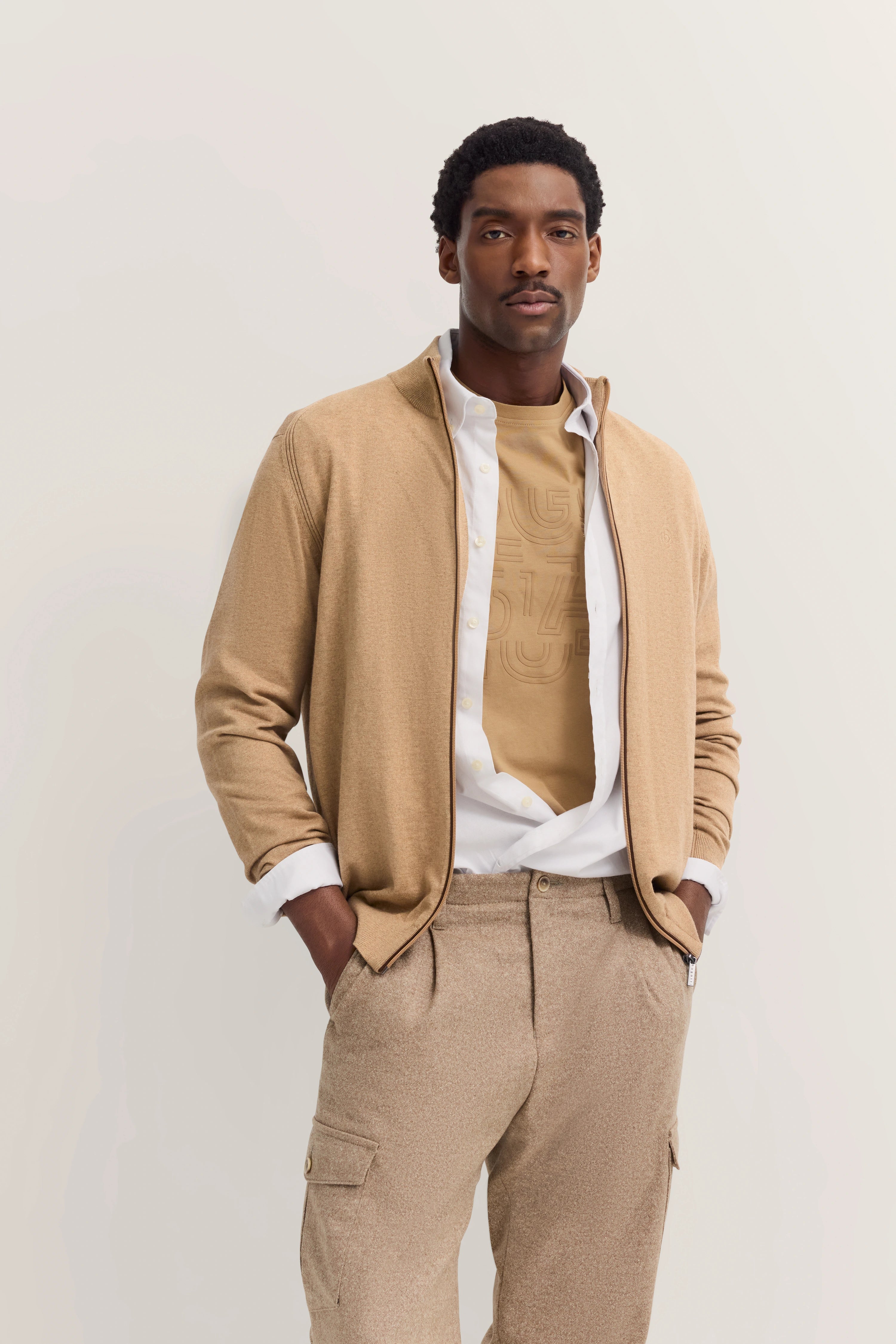 Model in beige bugatti-outfit: blazer, shirt, broek. Ontspannen pose, handen in zakken.