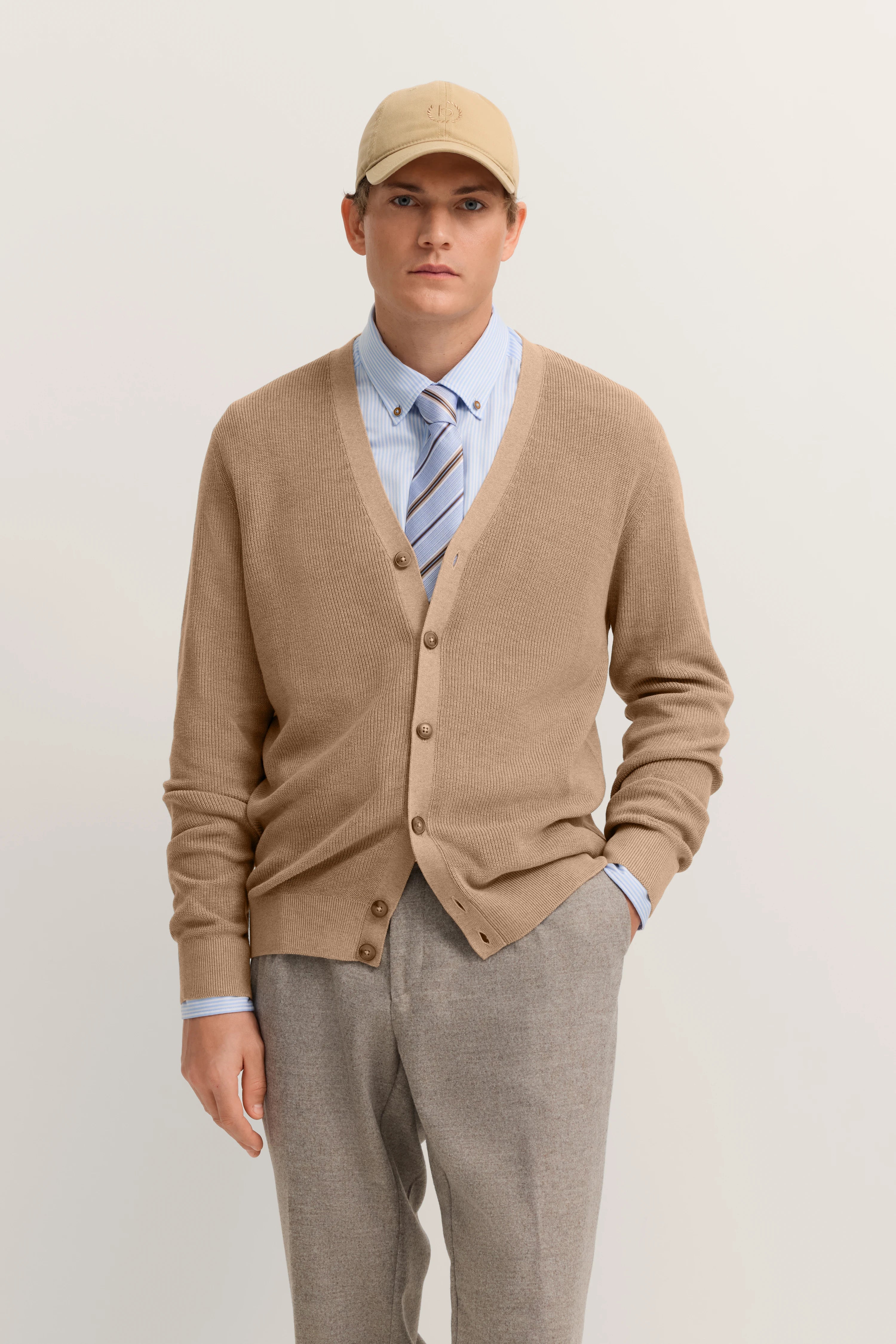 Beiger Cardigan, blaues Hemd, gestreifte Krawatte, graue Hose, beiges Cap, Model stehend, bugatti Stil.