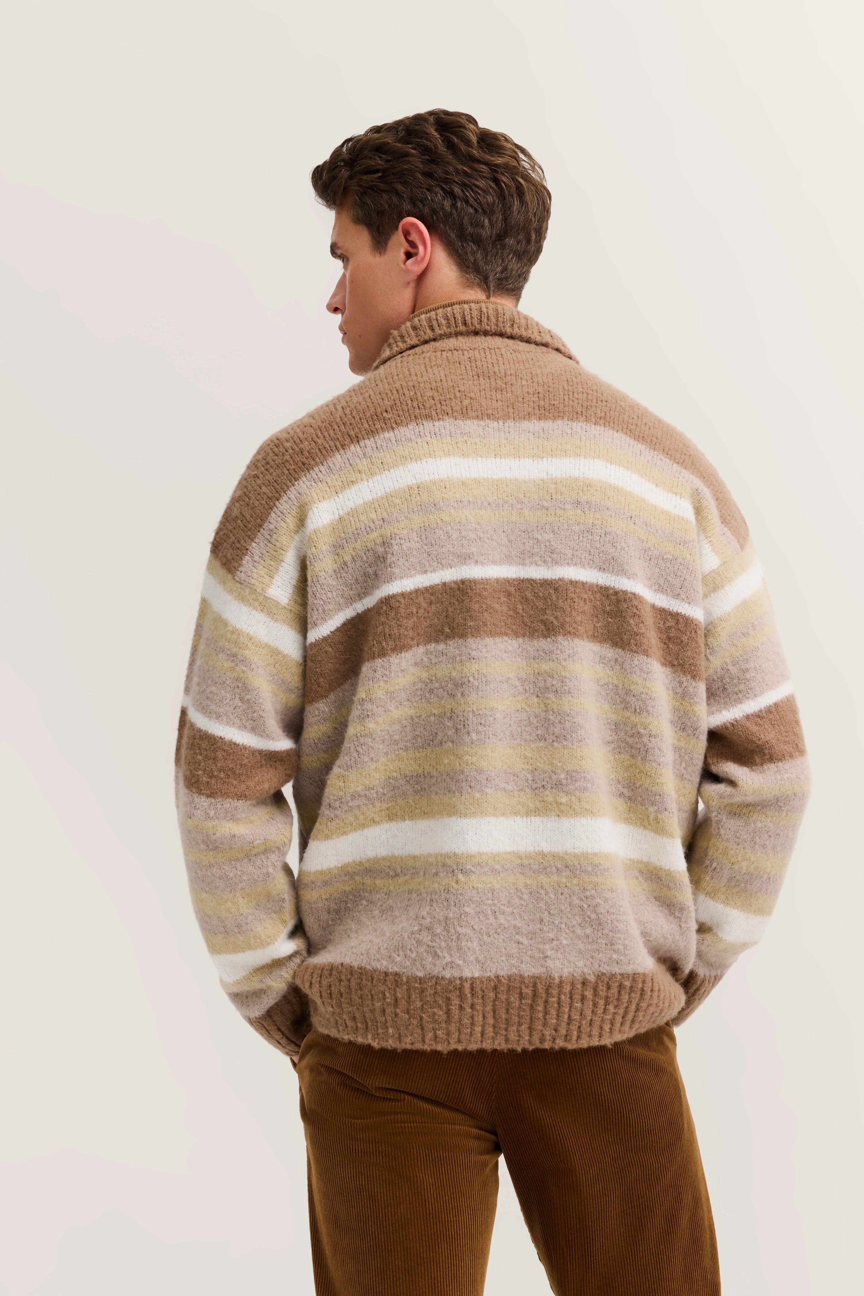 Model wearing striped beige and brown sweater, back view, paired with brown corduroy pants.