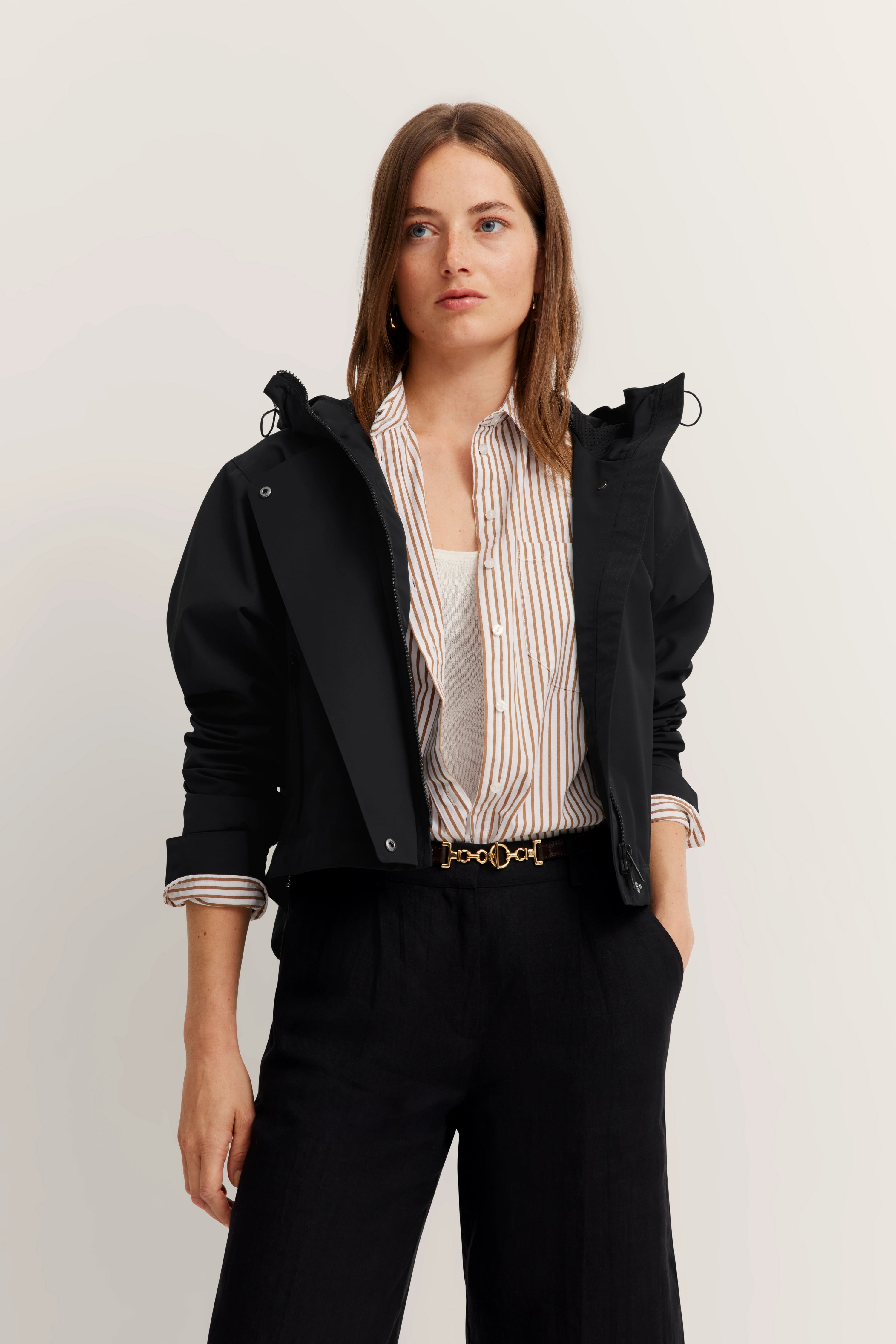 Model, vrouw in zwarte jas, gestreept shirt en zwarte broek, ontspannen pose.