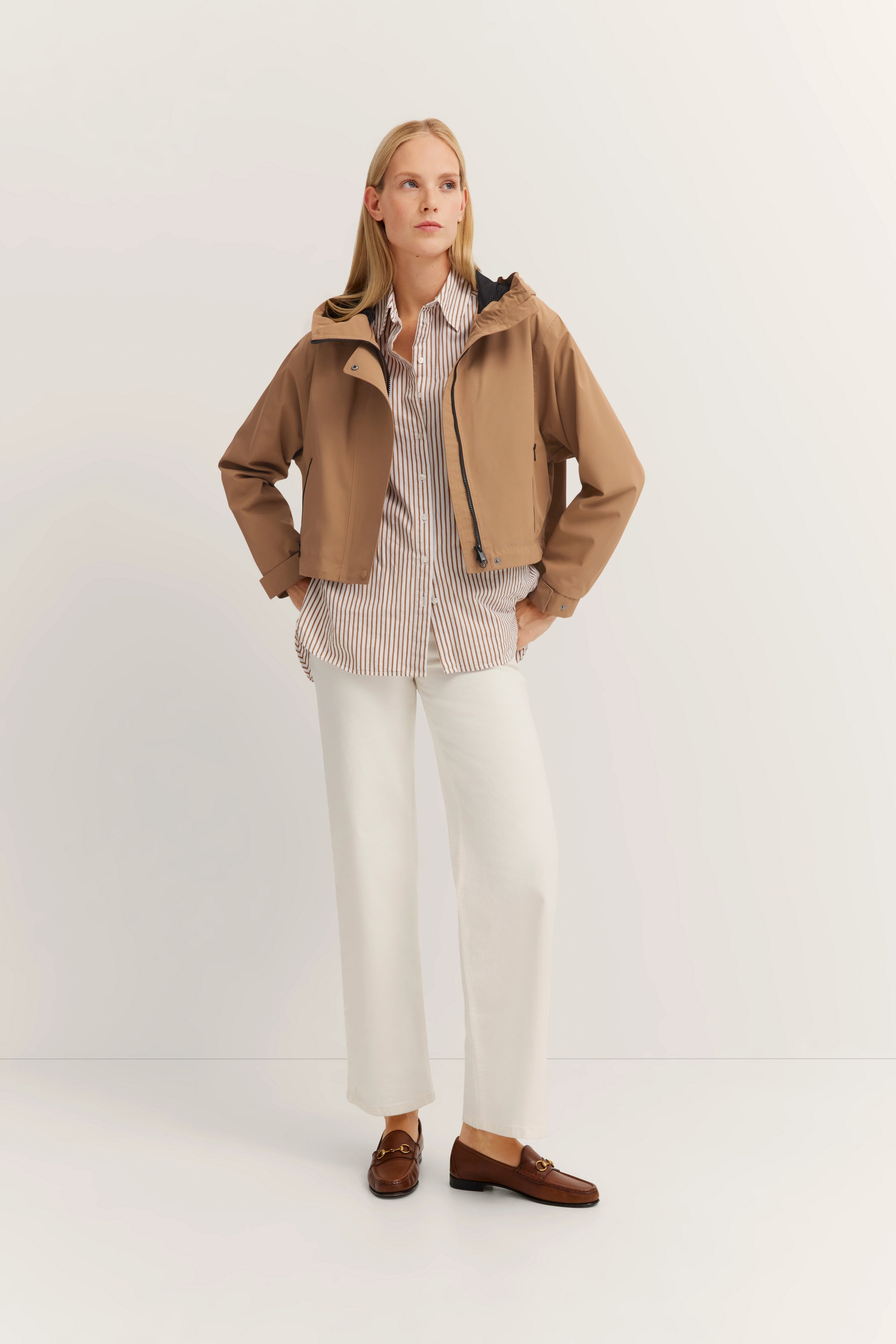 Tan Jacke, gestreiftes Hemd, weiße Hose, braune Loafer. Model posiert mit entspanntem Blick.