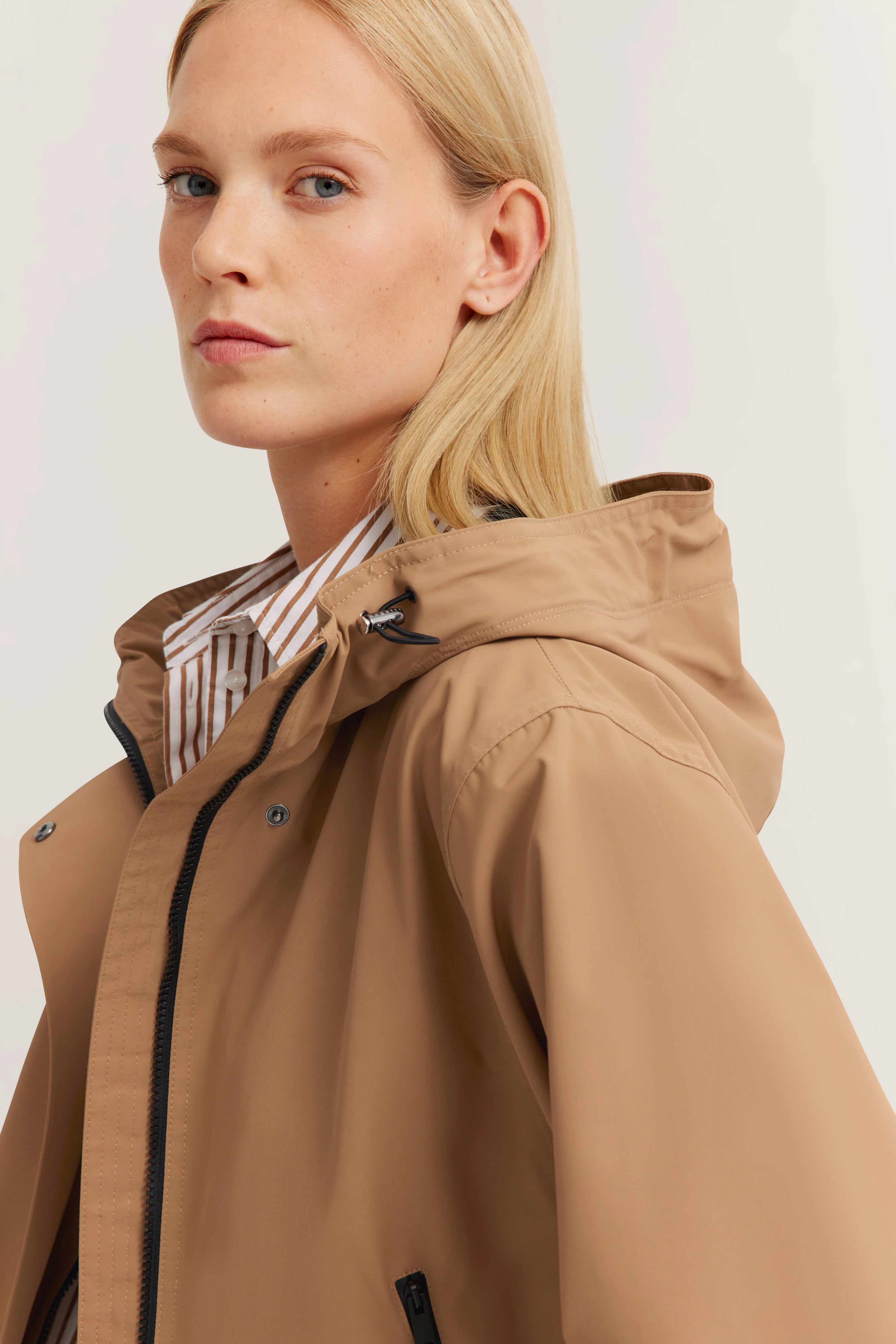Beige bugatti Regenjacke mit Kapuze, Modelbild, seitliches Portrait, gestreiftes Hemd im Hintergrund.