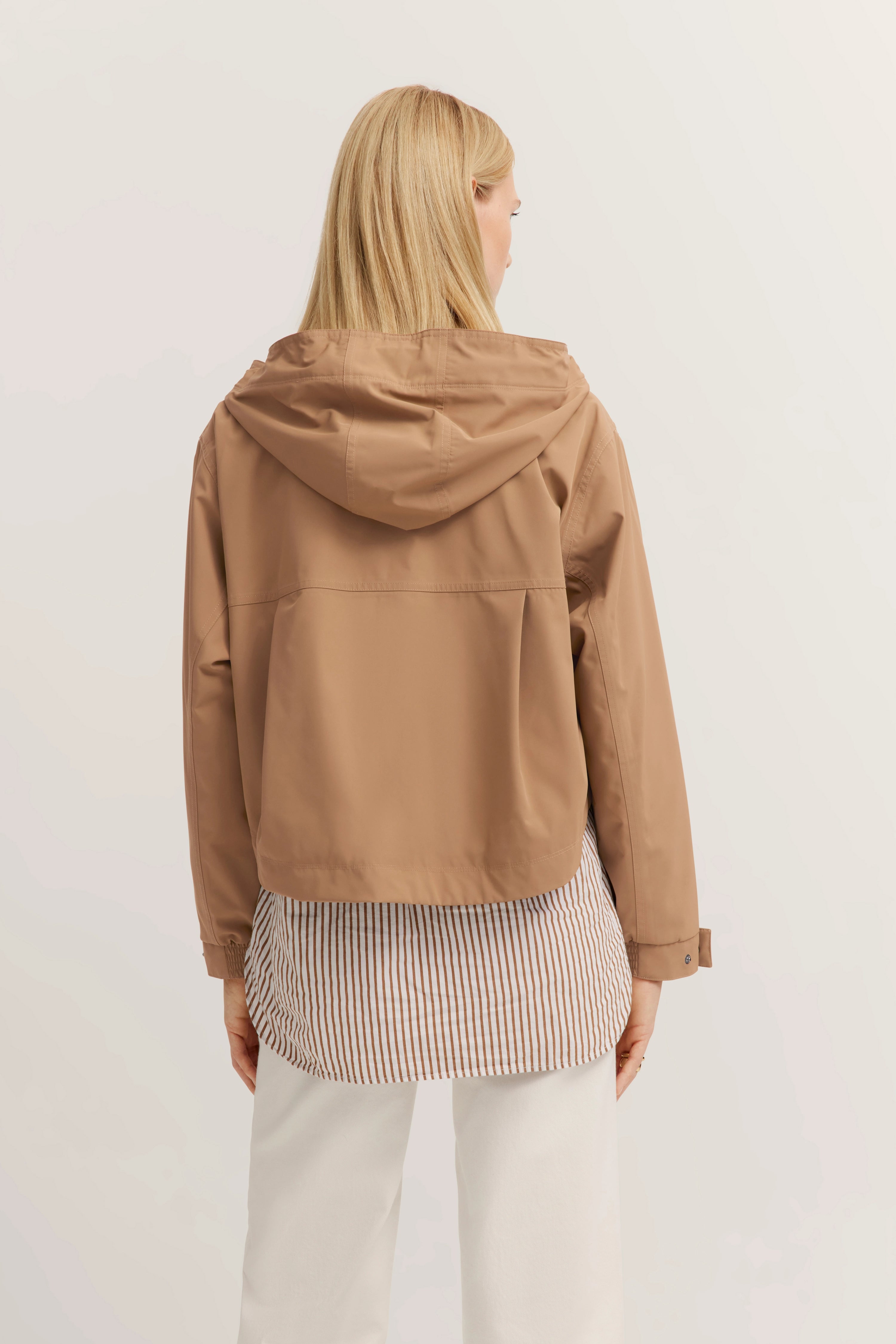 Blonde Person von hinten in camel Jacke, gestreiftem Hemd und weißen Hosen. Lässiges Modelbild.