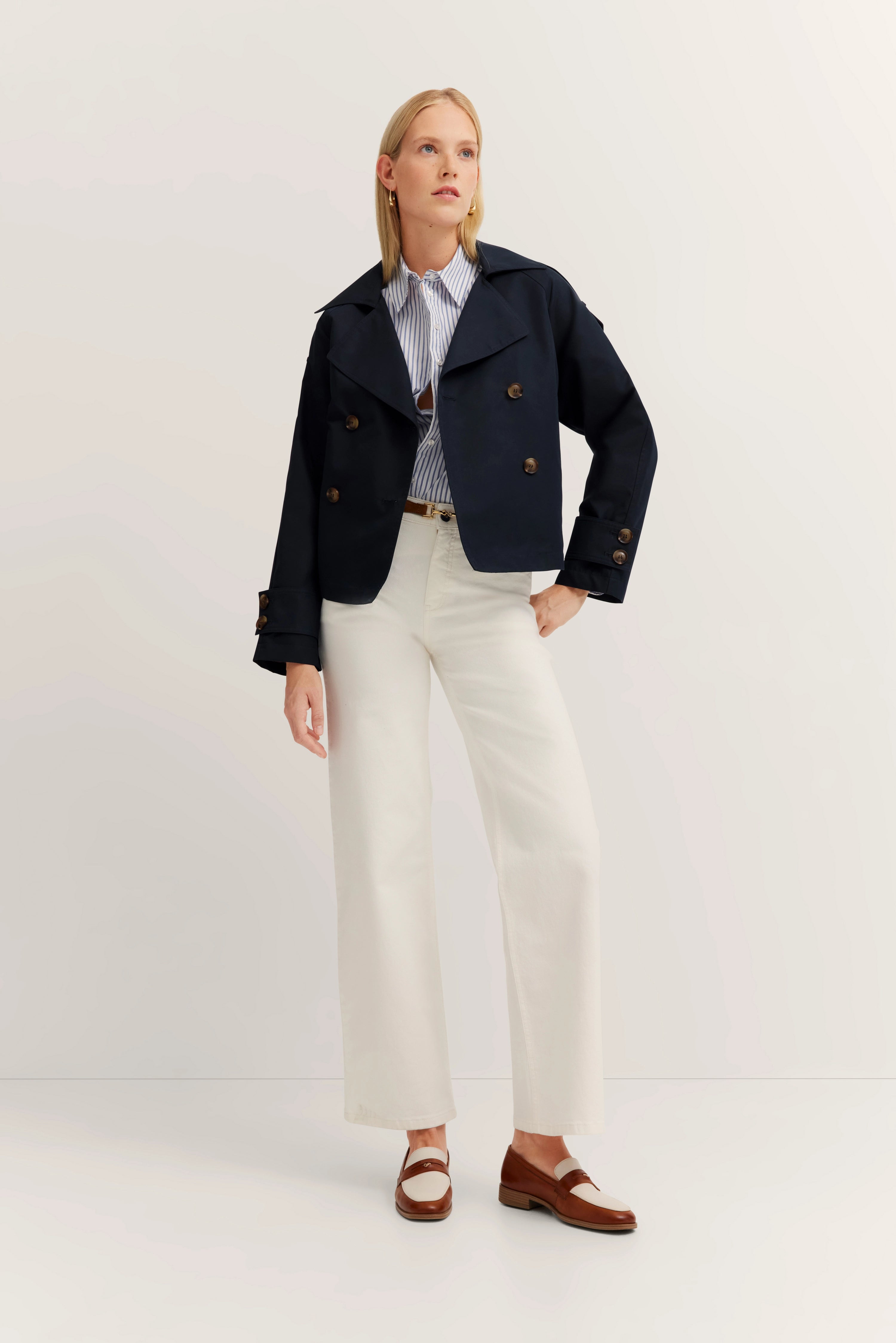 Blaues bugatti-Jackett, gestreifte Bluse, weiße Hose, braune Schuhe, legeres Modelbild, entspannte Pose.