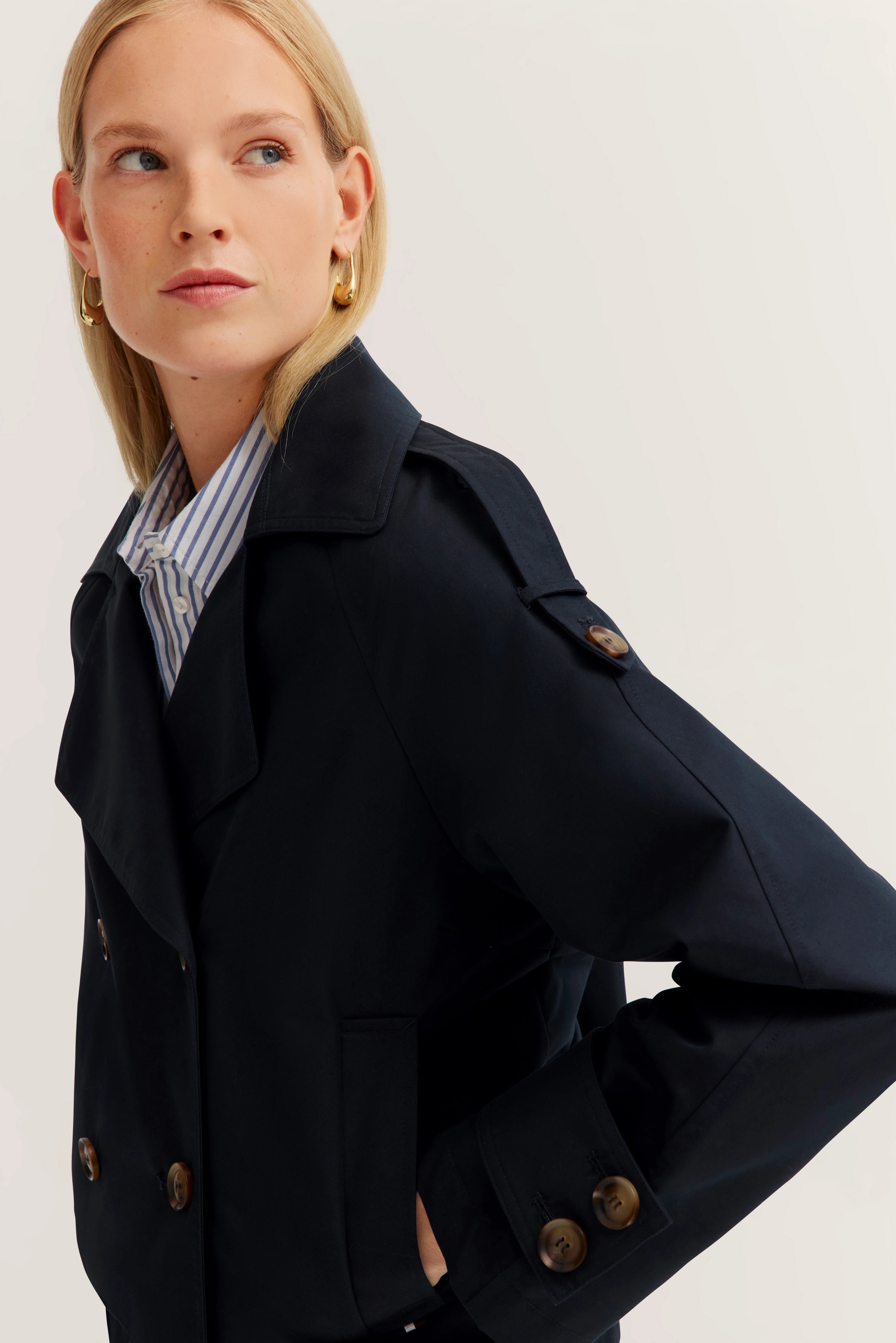Borstbeeld: Vrouw in donkerblauwe trenchcoat, gestreept overhemd, gouden oorbellen.