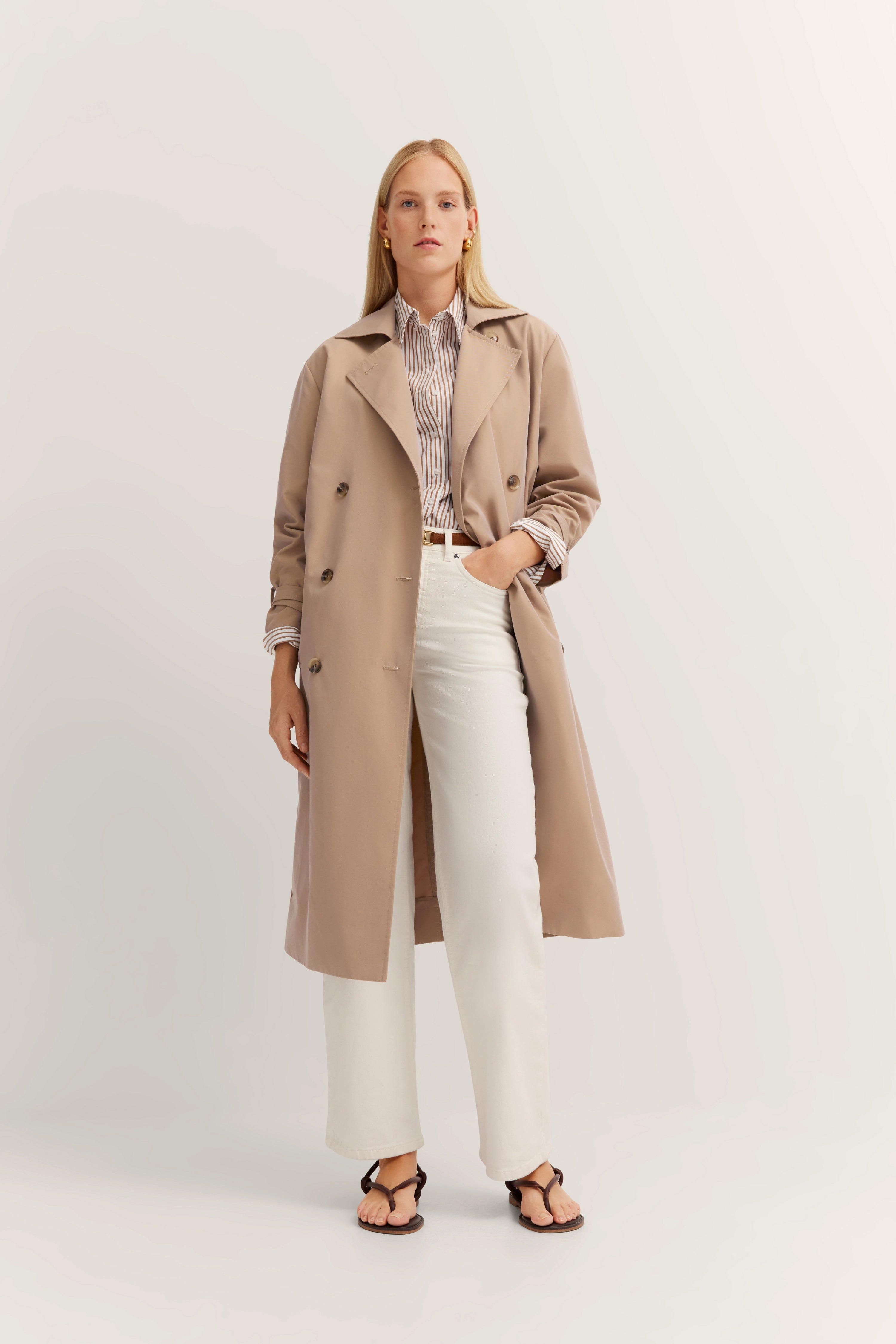 Beiger Trenchcoat, gestreiftes Hemd, weiße Hose. Modelbild, lässiges Stehen mit Hand in der Tasche.