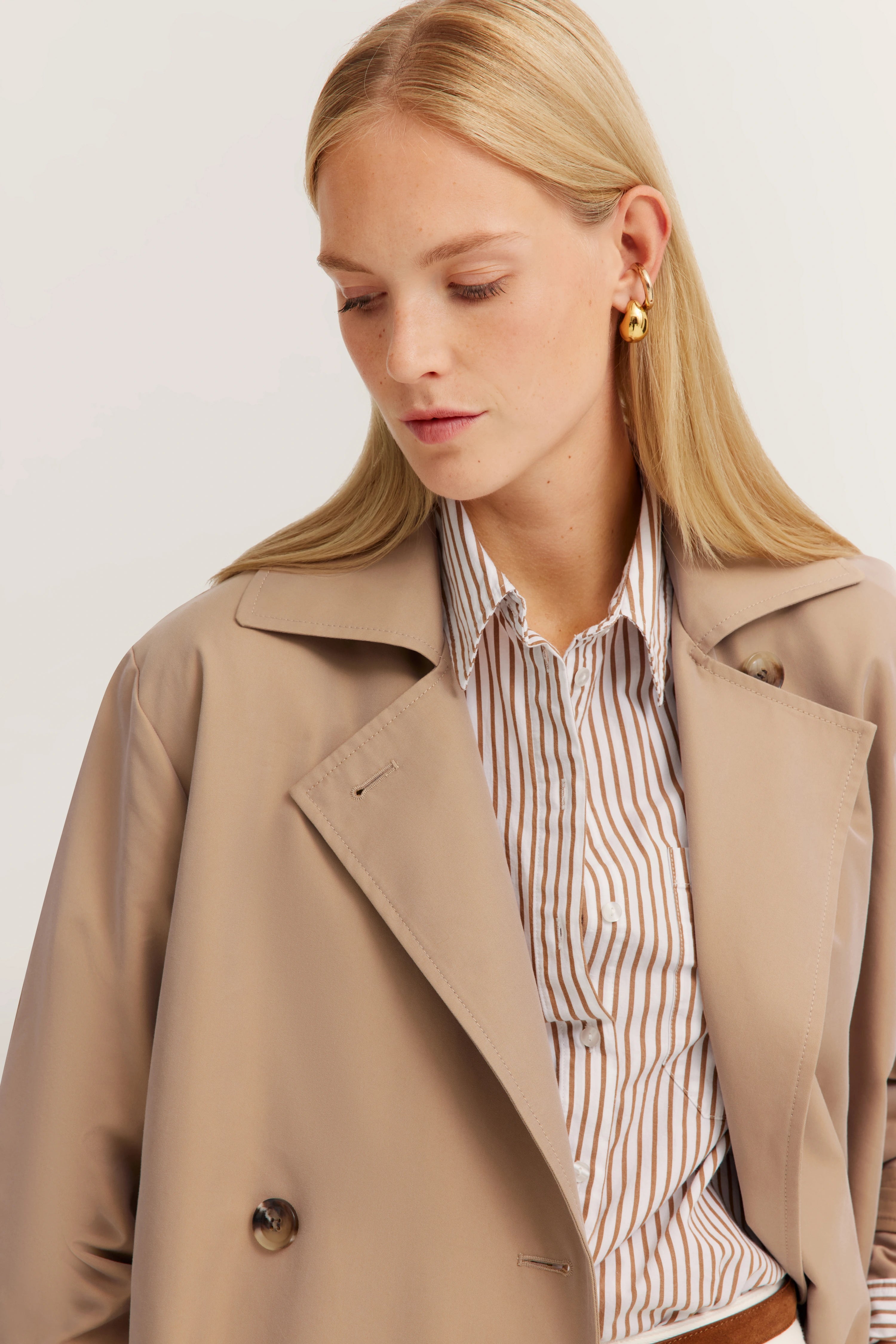 Beigefarbener Trenchcoat über gestreifter Bluse, Modelbild mit seitlichem Blick.