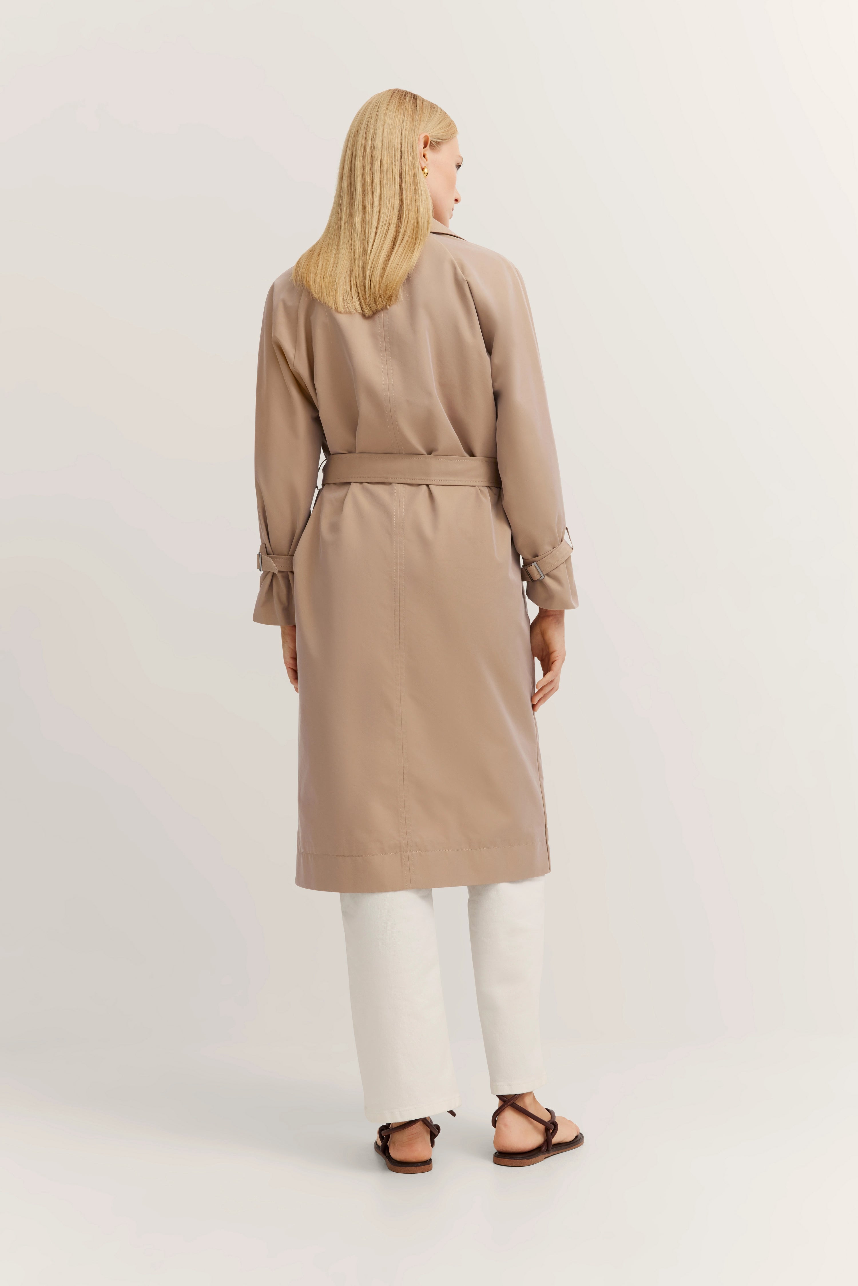 Beige trenchcoat van bugatti, achteraanzicht, boven witte broek, model staat ontspannen.