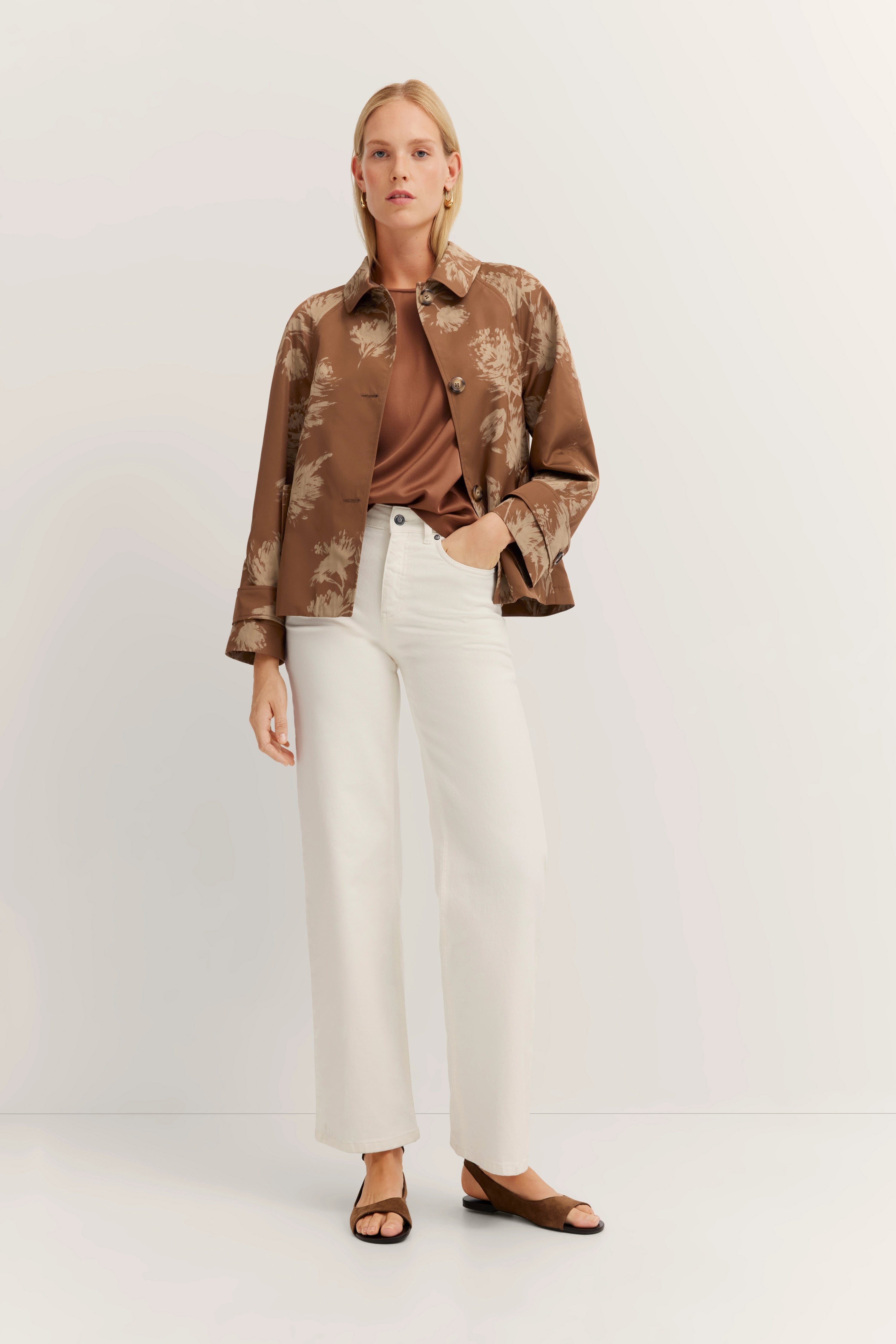 Braune Jacke mit Muster, braunes Shirt, weiße Hose. Model posiert stehend.