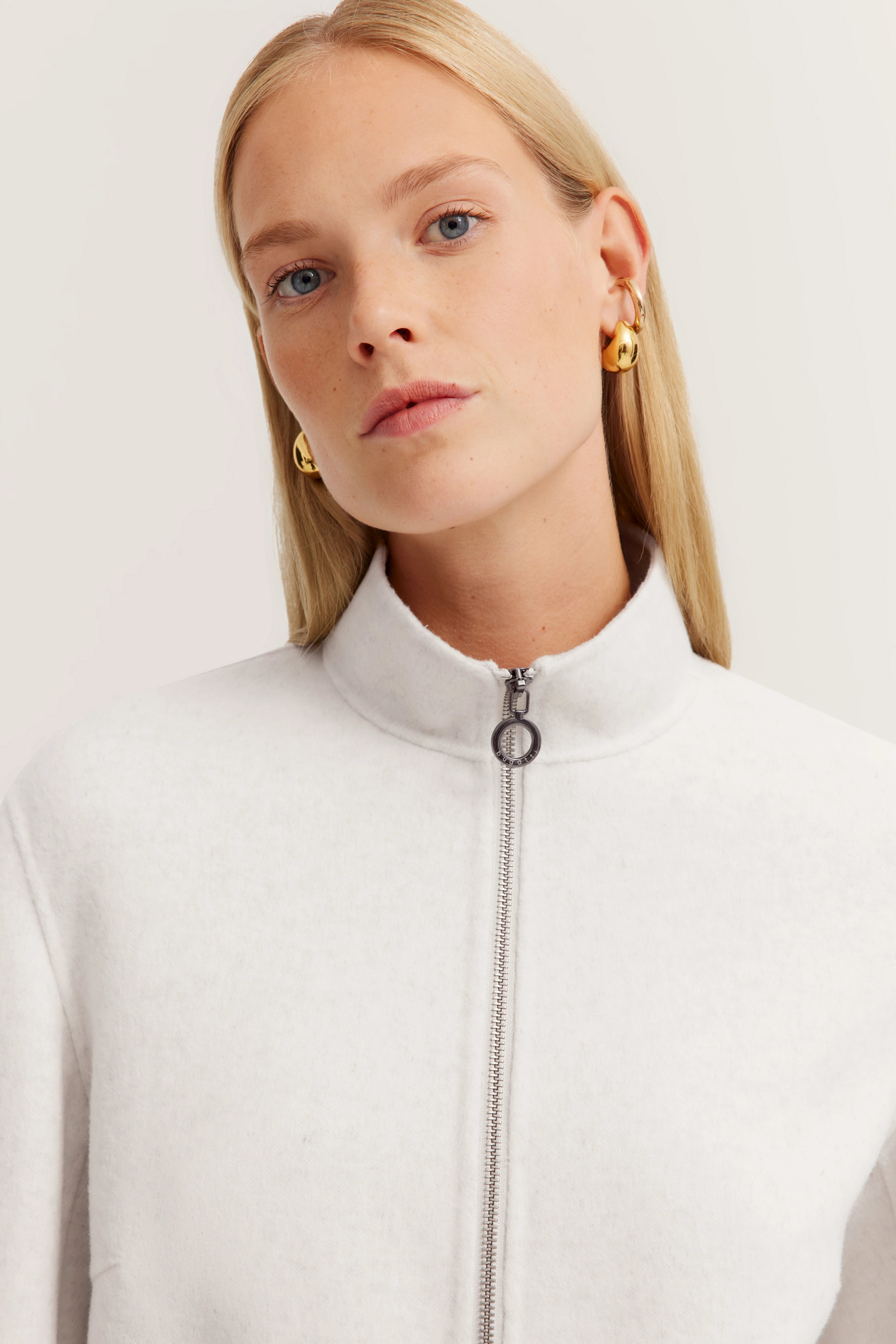 Büstenbild von Model in weißem bugatti-Oberteil mit Reißverschluss und goldenen Ohrringen.