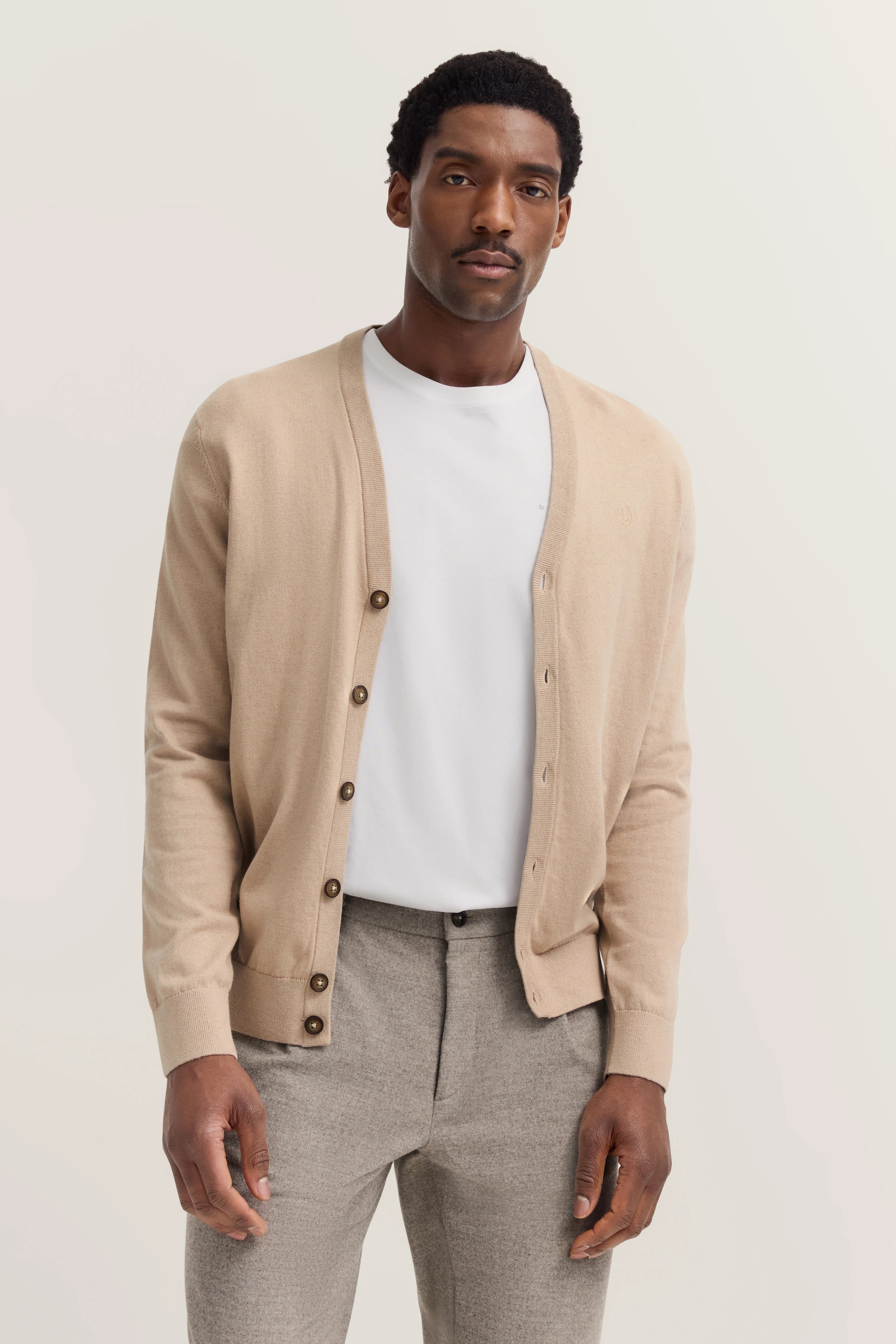 Beiger bugatti Cardigan, weißes Shirt, graue Hose, legeres Modelbild, entspanntes Posing.