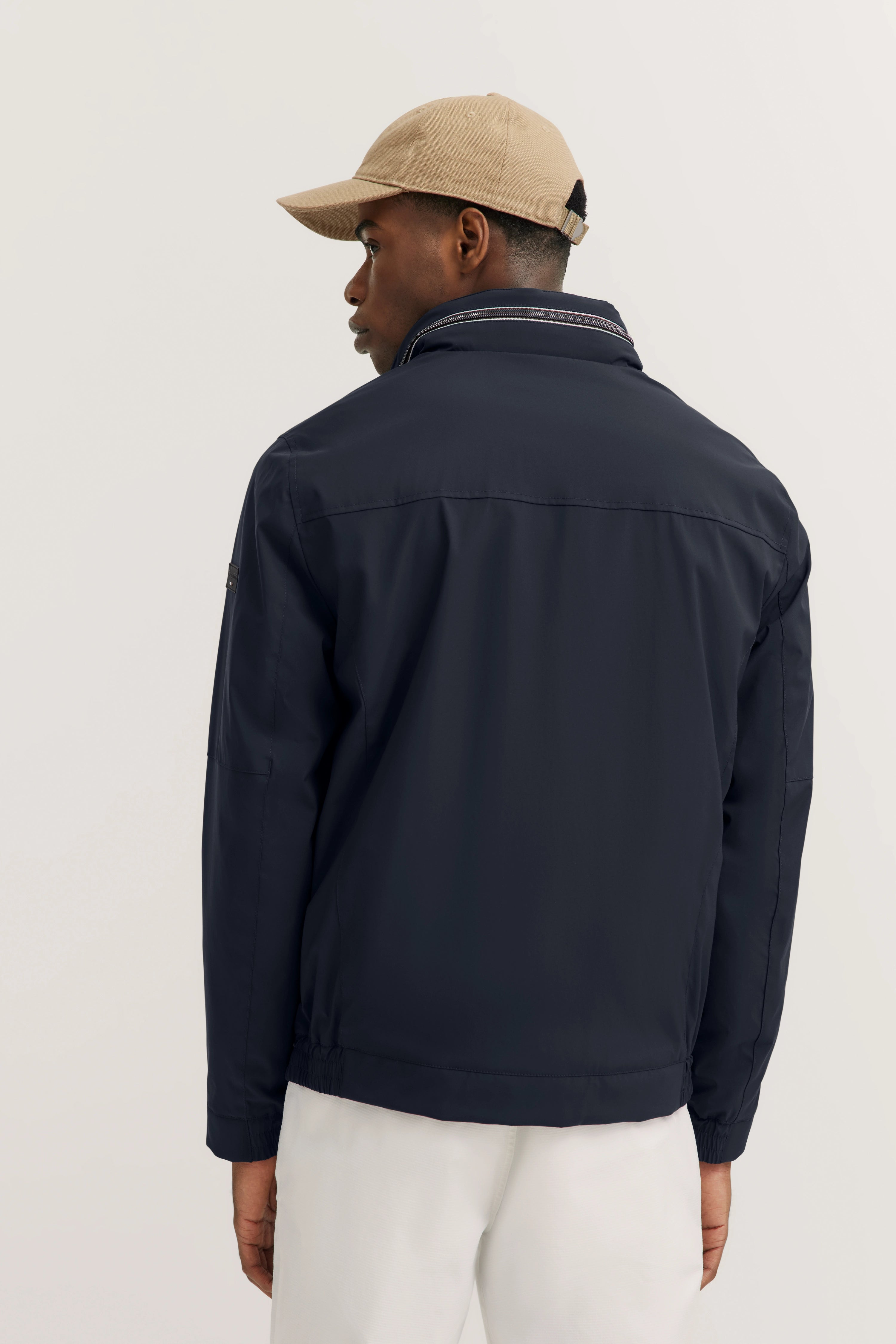 Dunkelblaue bugatti Jacke, Model von hinten, beige Mütze, weiße Hose, entspannte Haltung.