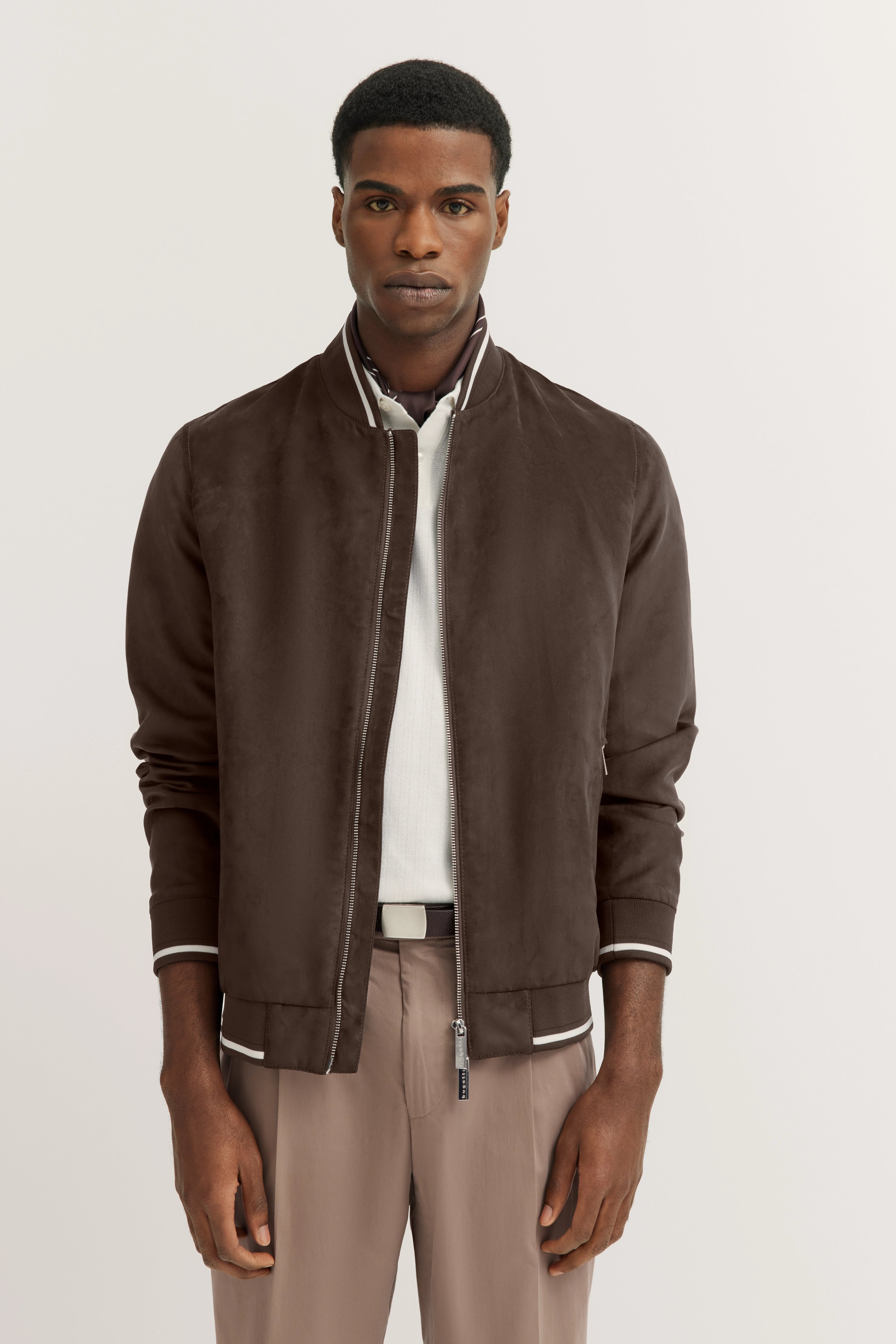 Model in brauner bugatti Bomberjacke, weißem Polo, beigen Hosen. Stehpose, klassisch-elegant.