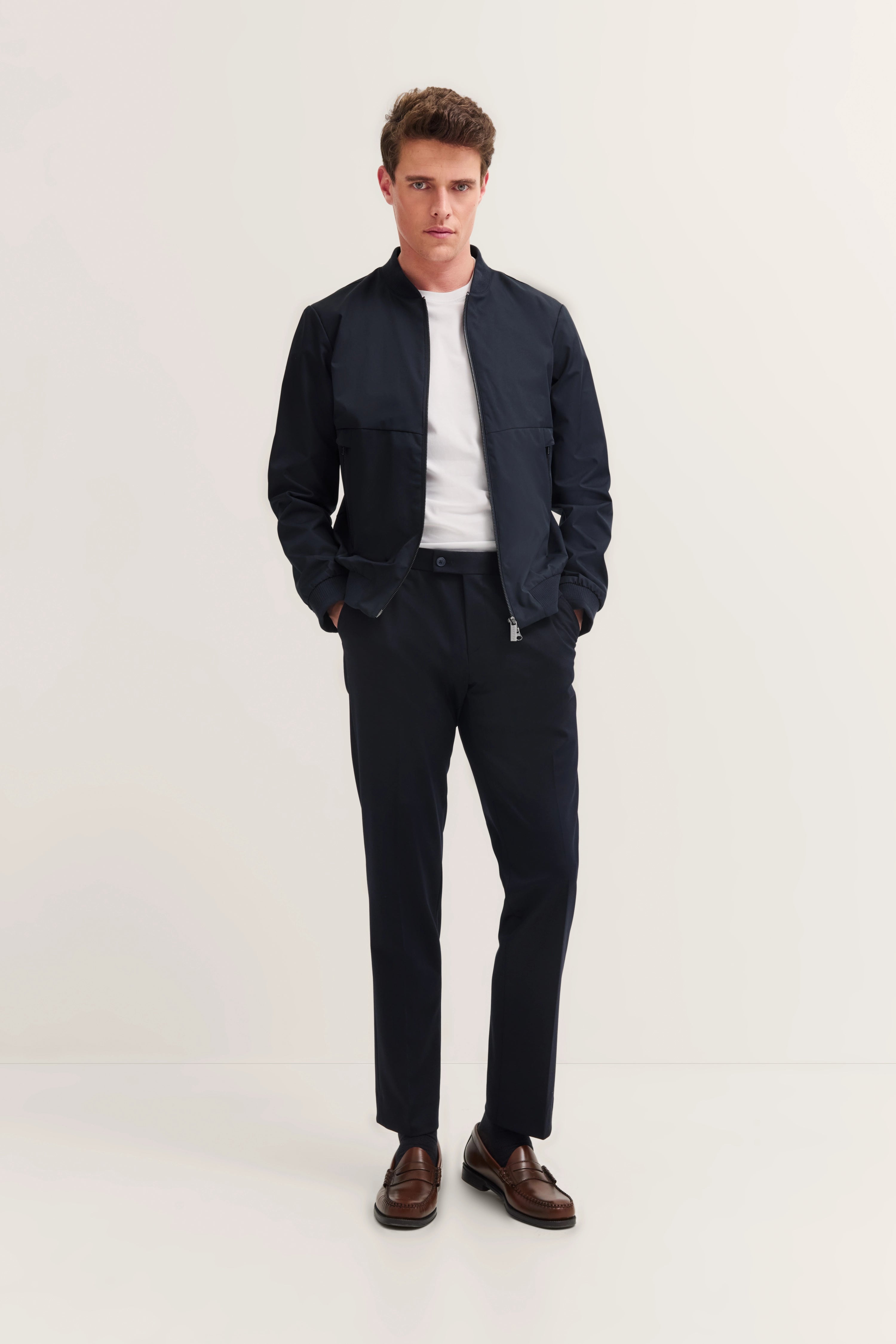 Model in schwarzer bugatti Jacke, weißem Shirt und Hose, braune Schuhe, lässige Pose, Hände in Taschen.