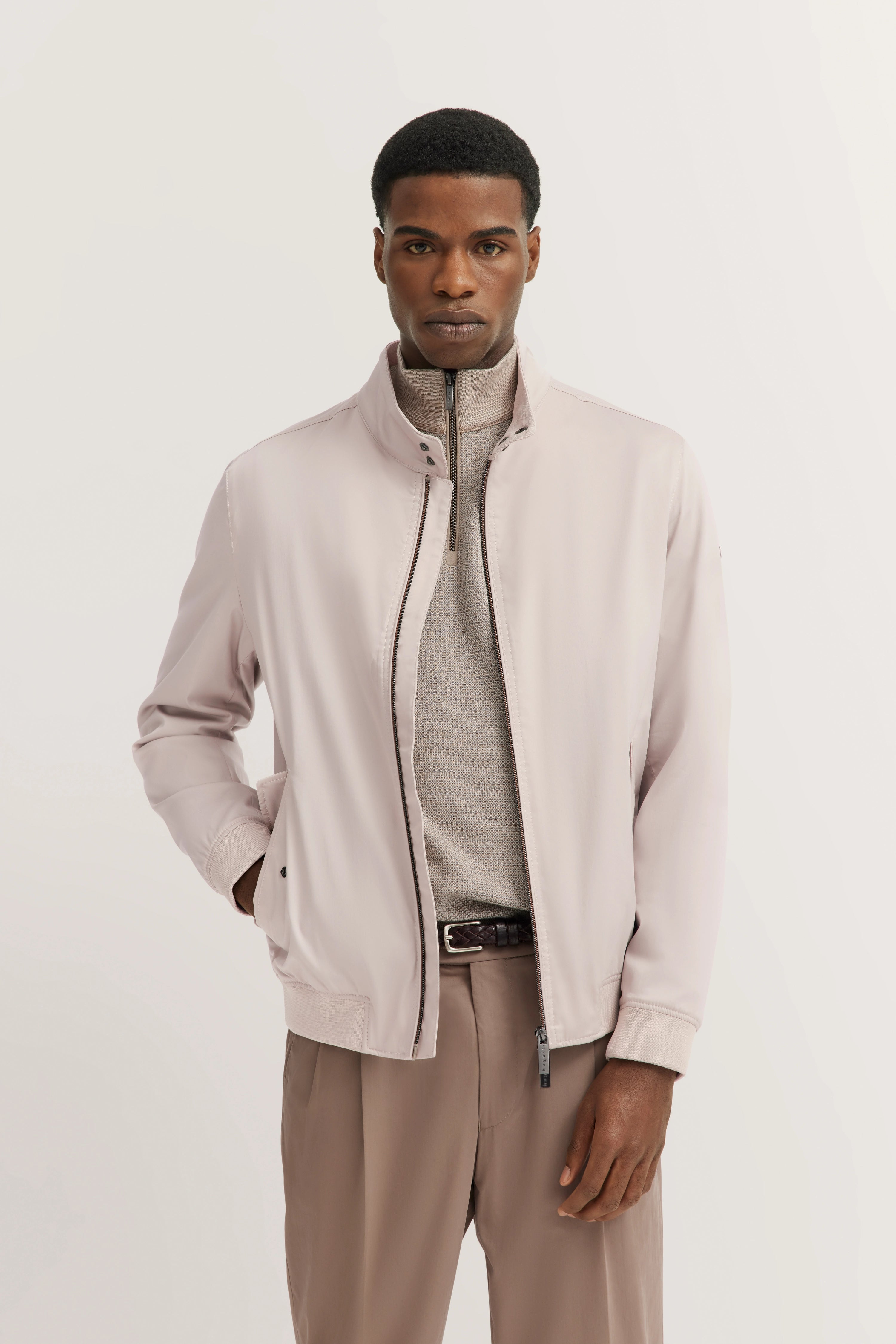 bugatti Modelbild: Beige Jacke, grauer Pullover, braune Hose, entspannte Pose, Hände in Taschen.