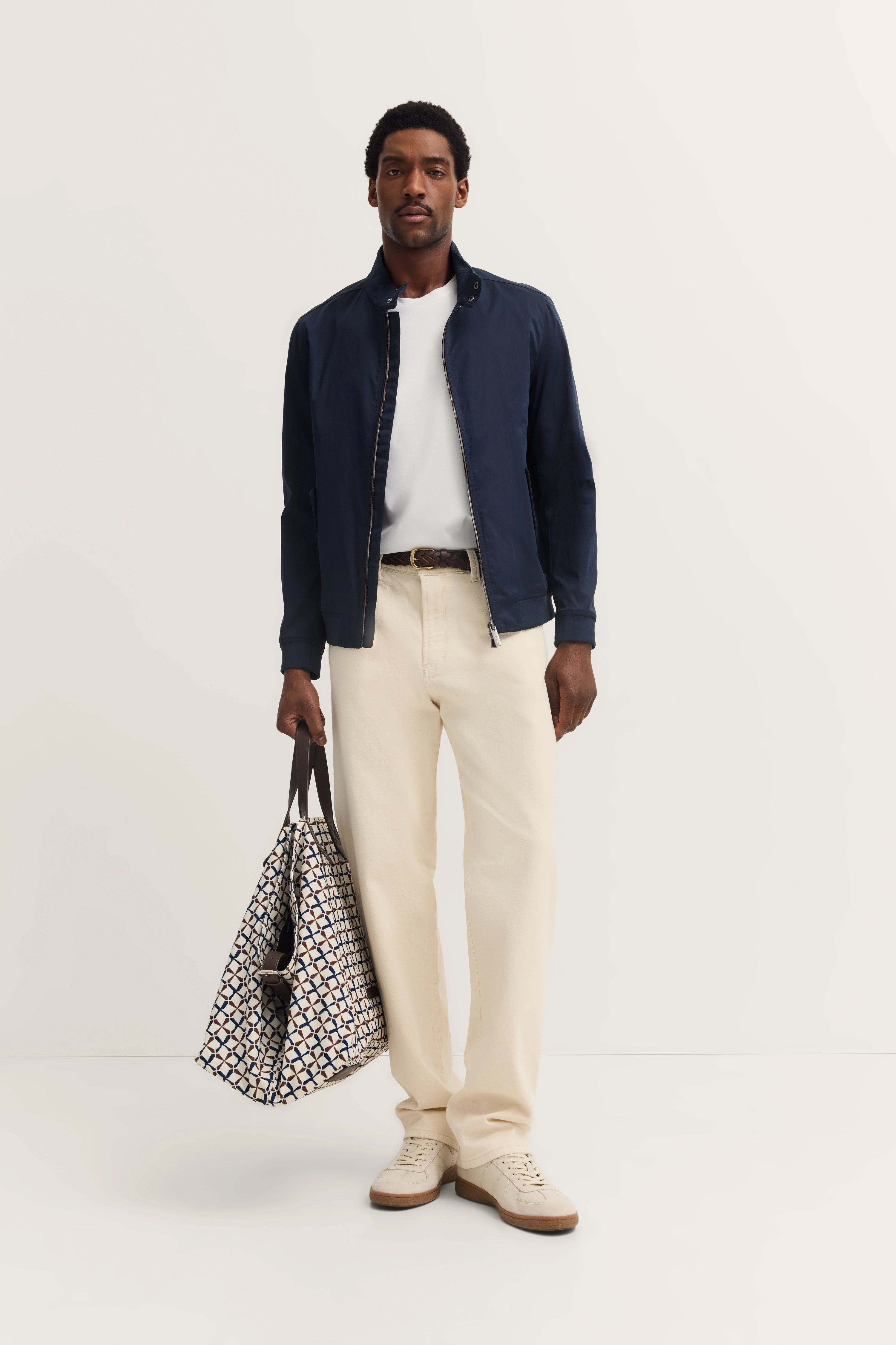 Model in navy jacket, white shirt, cream pants. Geometric bag, relaxed pose.