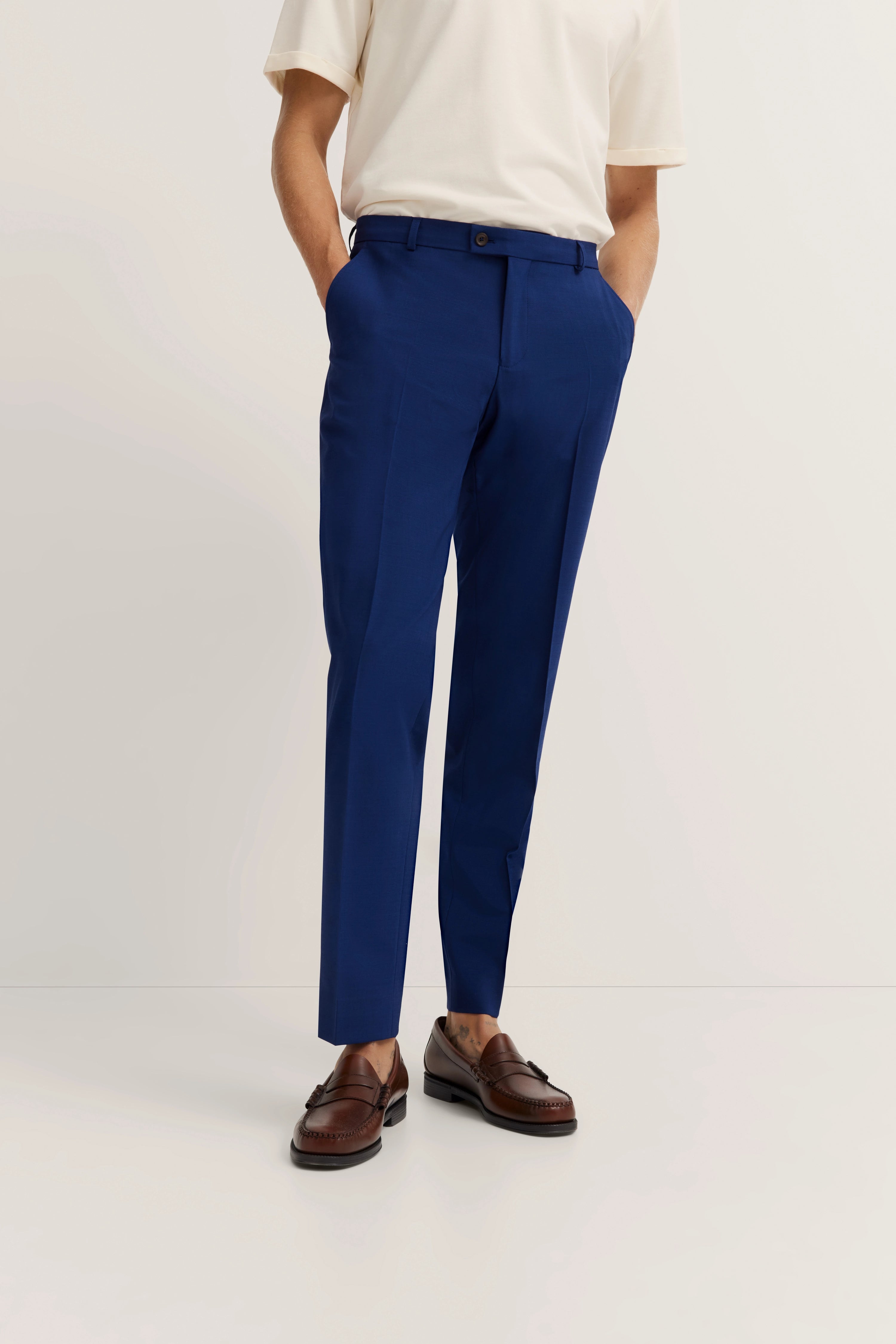 Herrenoutfit mit cremefarbenem Shirt, blauer Hose, braunen Schuhen, Model lässig stehend.