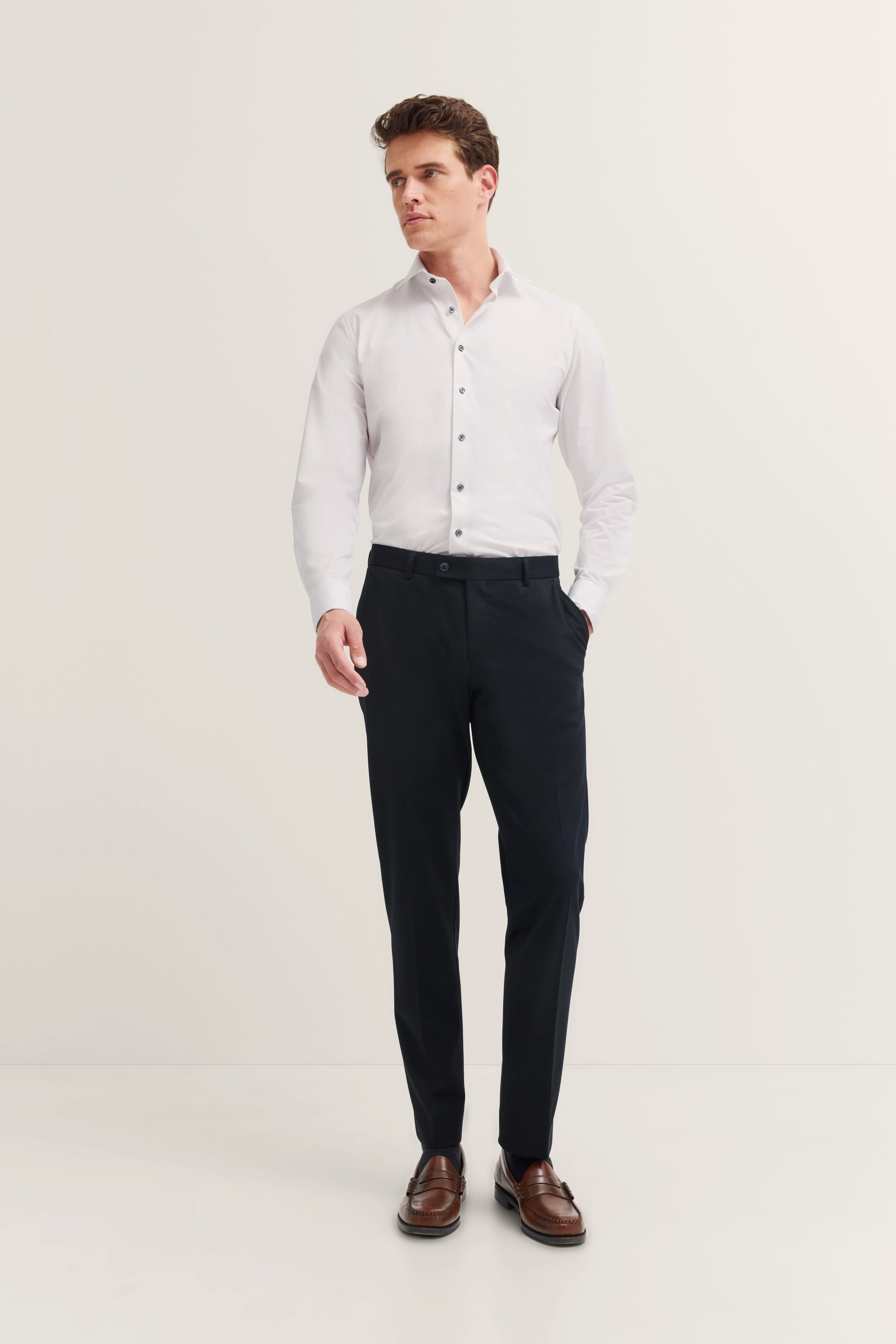 Model in weißem Hemd und dunkelblauer Hose, stehend, mit einer Hand in der Tasche.