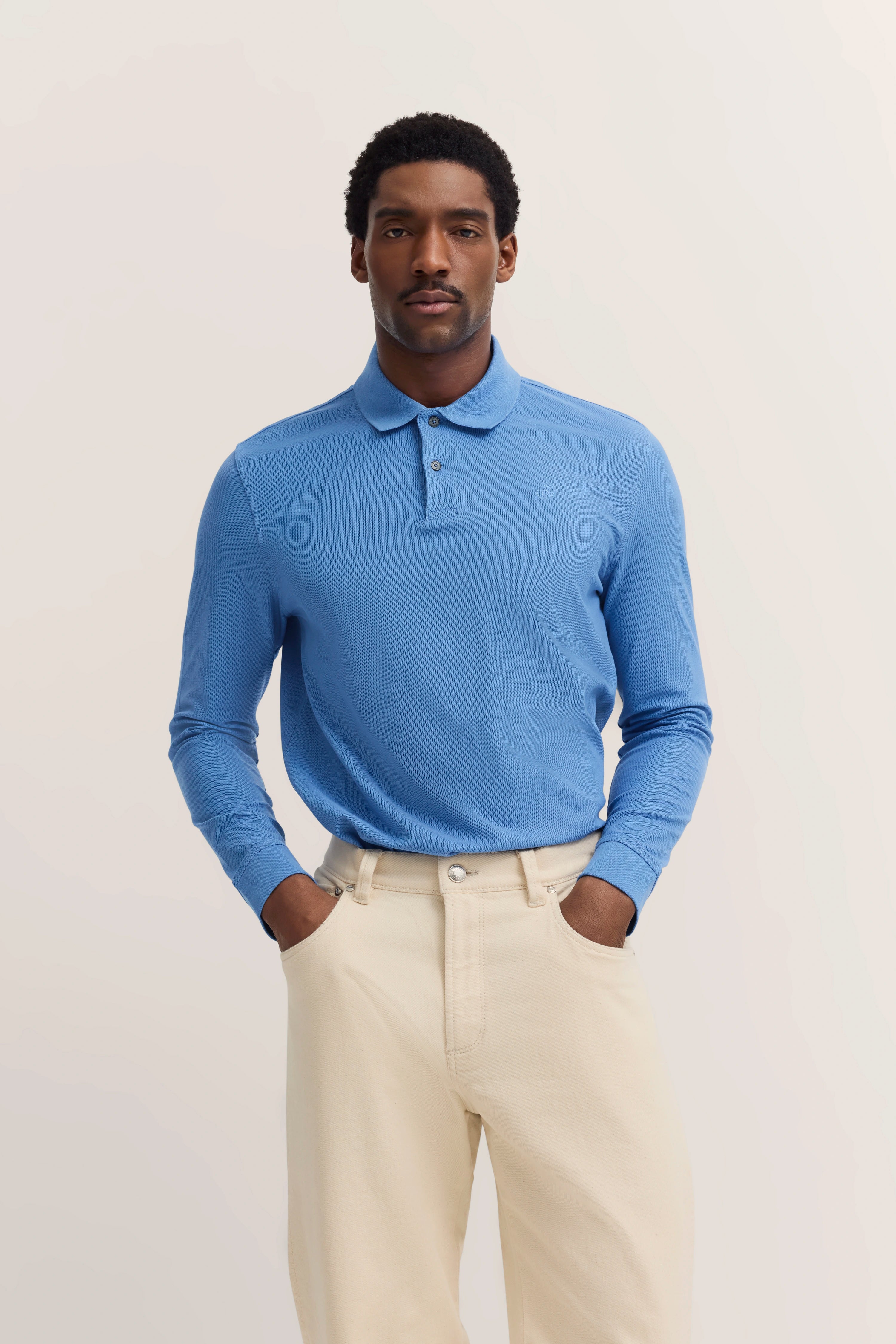Bugatti Modelbild: Blaues Poloshirt, beige Hose, entspannte Haltung, Hände in Hosentaschen.