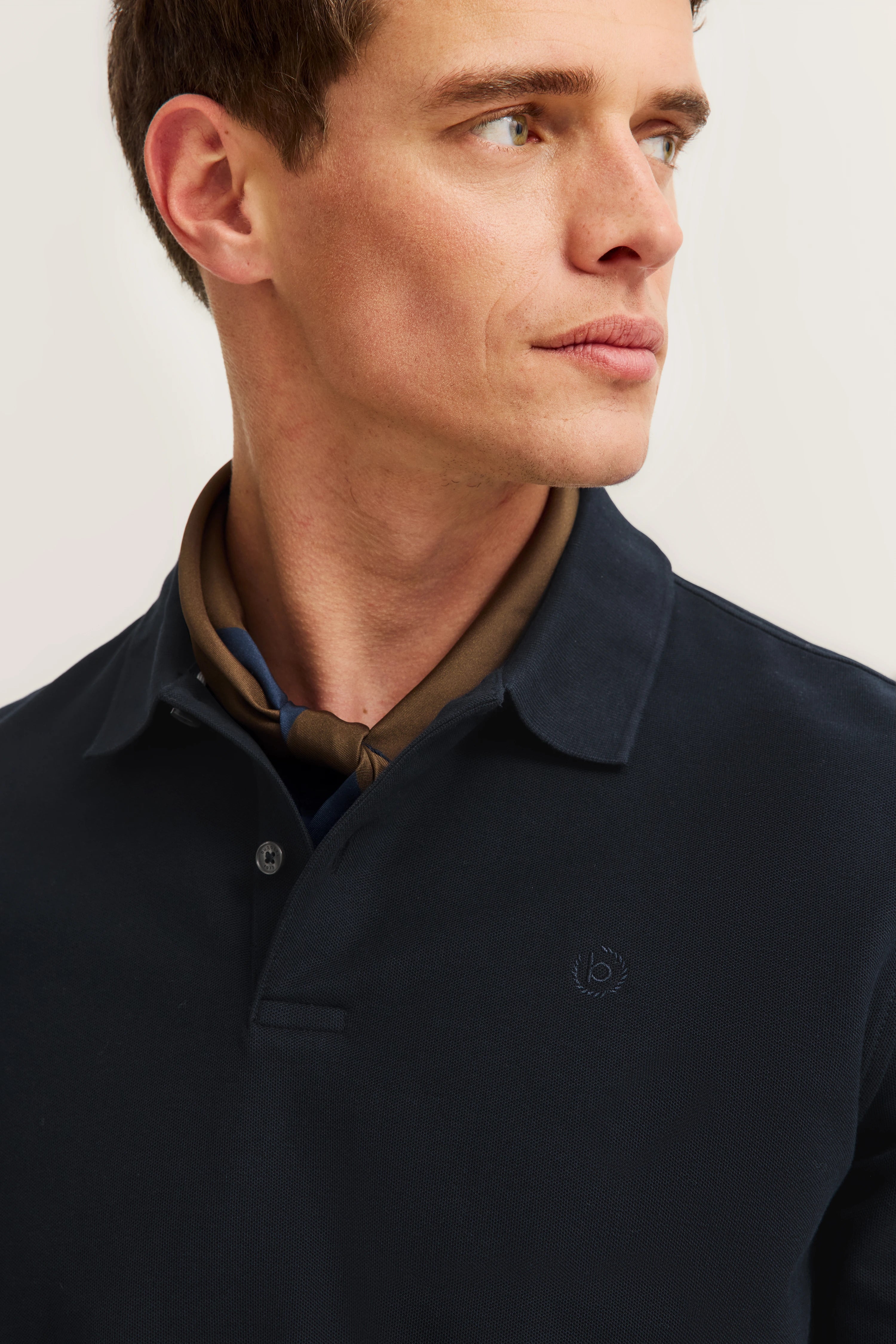 Model in dunkelblauem bugatti-Poloshirt mit braunem Schal, seitlich blickend. Eleganter, zeitloser Stil.