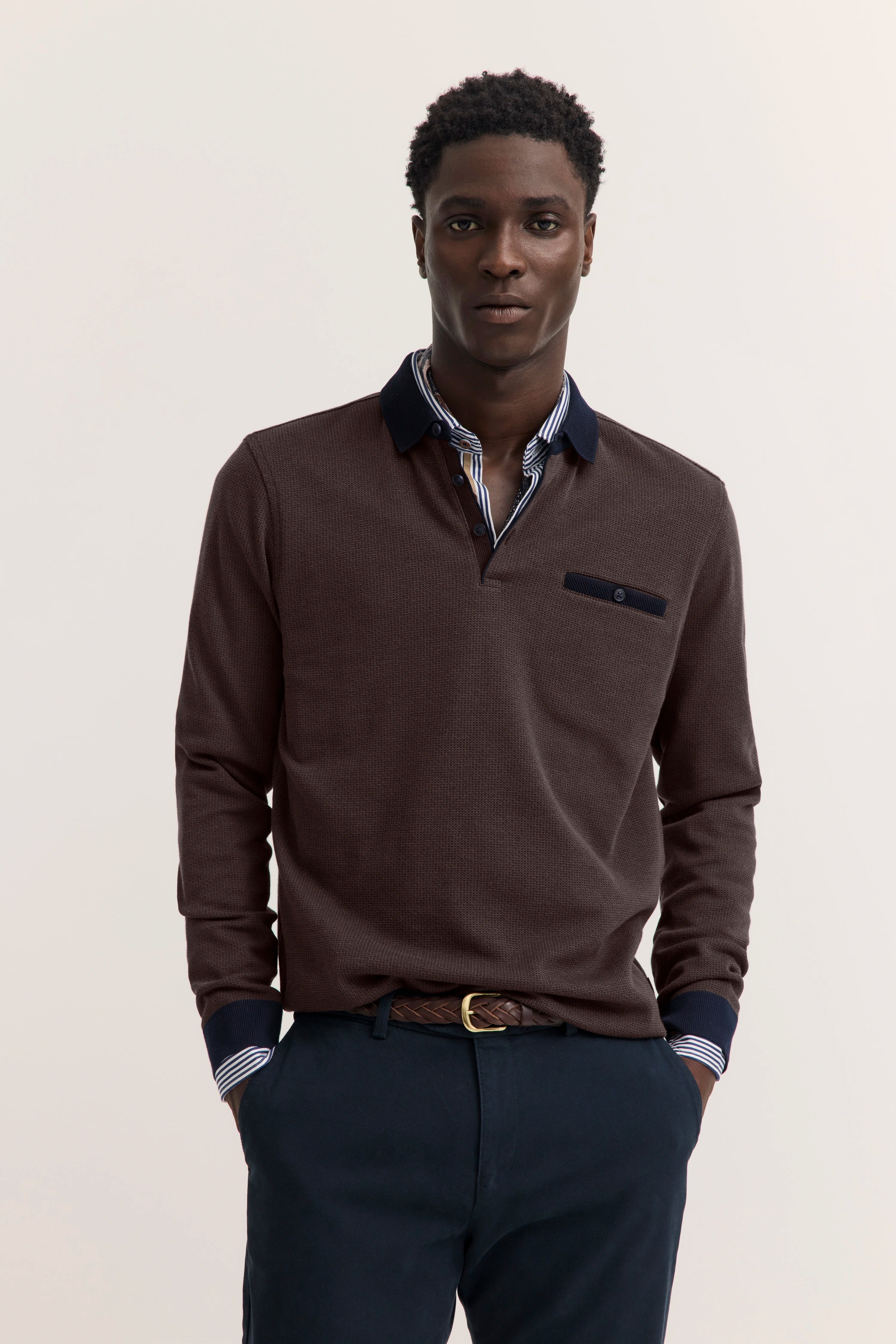 bugatti Modelbild: brauner Pullover, gestreiftes Hemdkragen-Detail, lässige Pose, Hände in Hosentaschen.