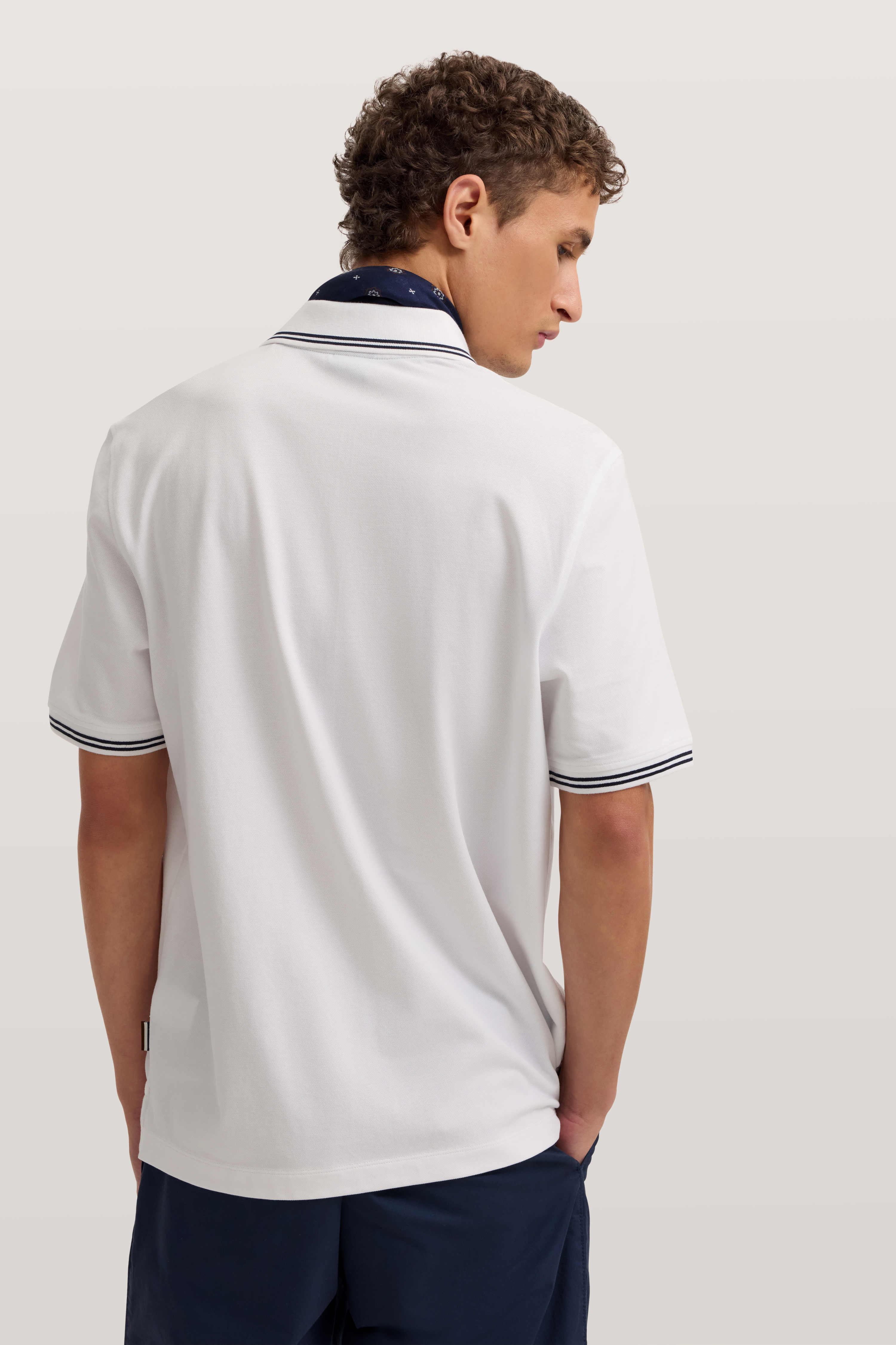 Weißes bugatti Polo, schwarzer Kragen, marineblaue Shorts. Model von hinten, entspanntes Posing.