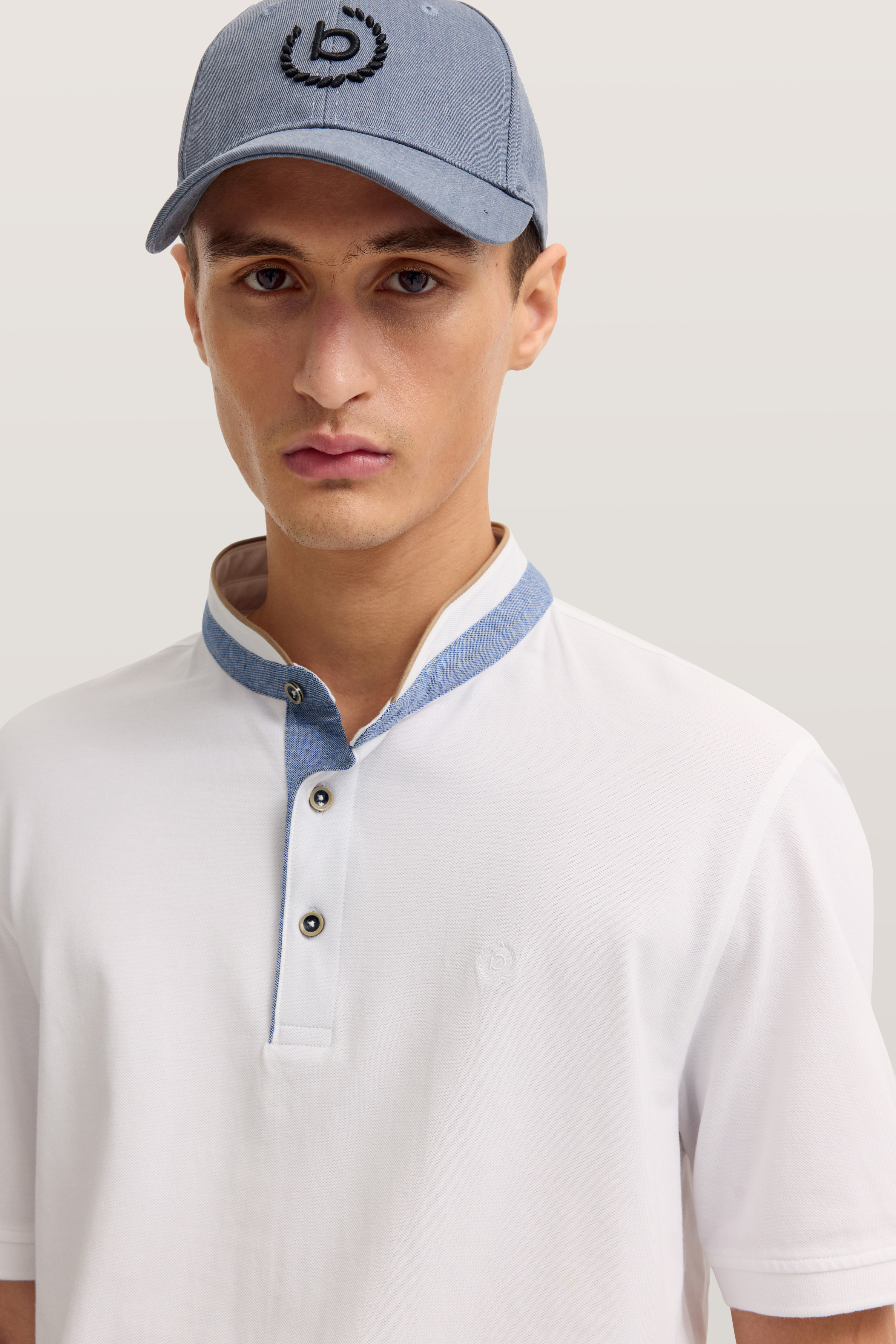 Weißes bugatti Polohemd mit blauem Kragen, Model mit blauer Mütze, frontal stehend.