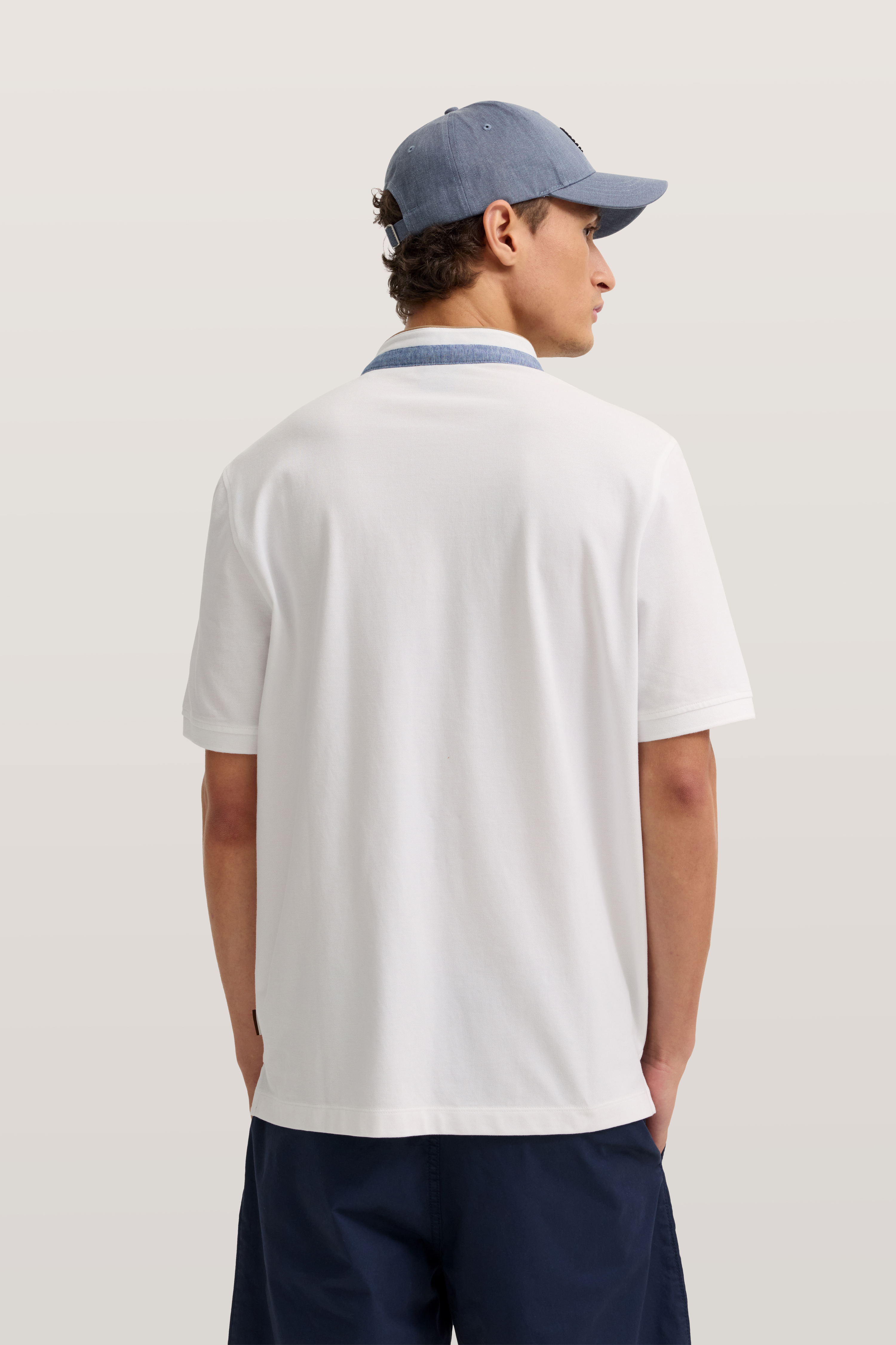 bugatti Modelbild: Weißes Polohemd, blau umrandeter Kragen, blaue Kappe, lässige Pose von hinten.