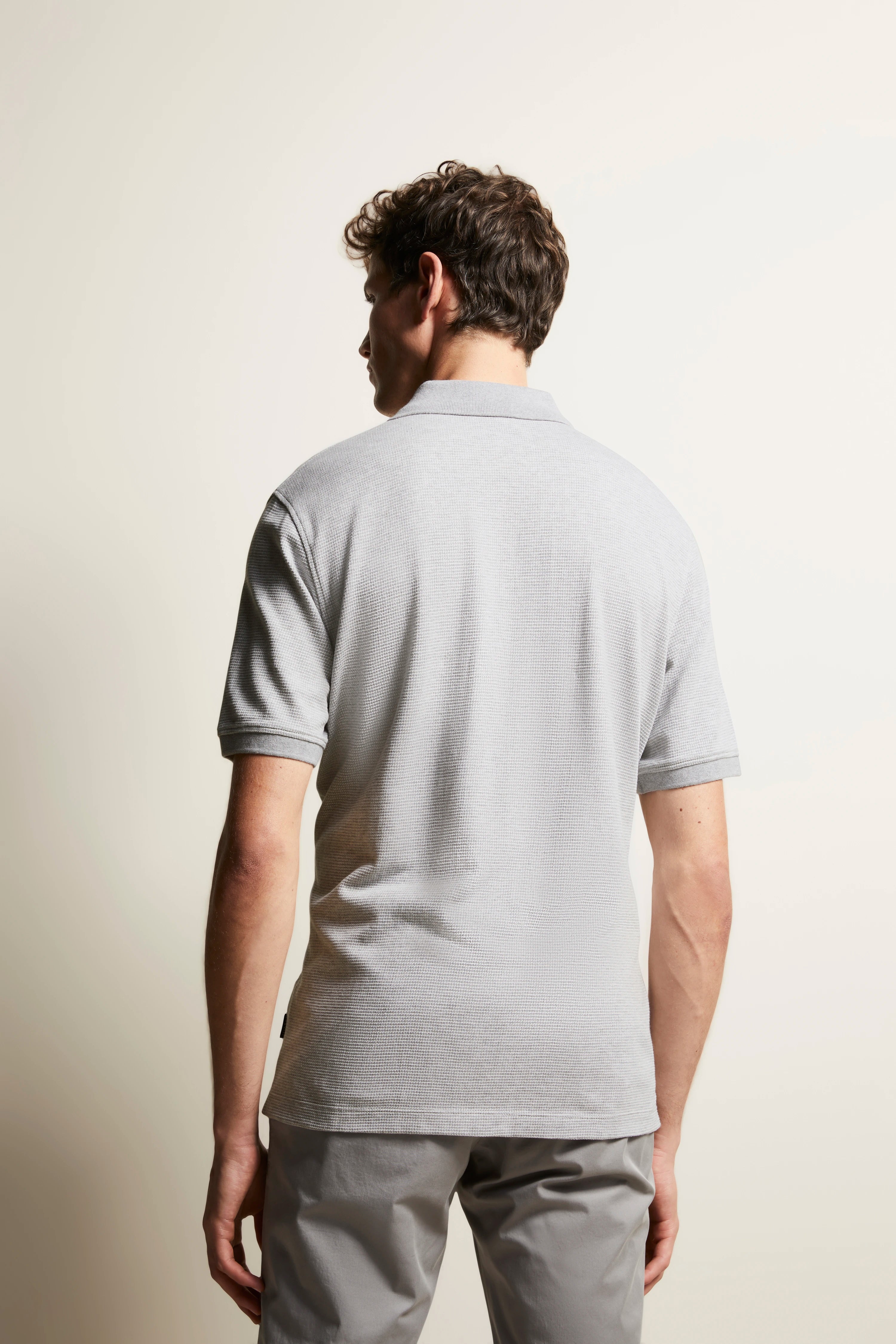 Gray polo shirt and beige pants, back view, model stands casually looking sideways.
