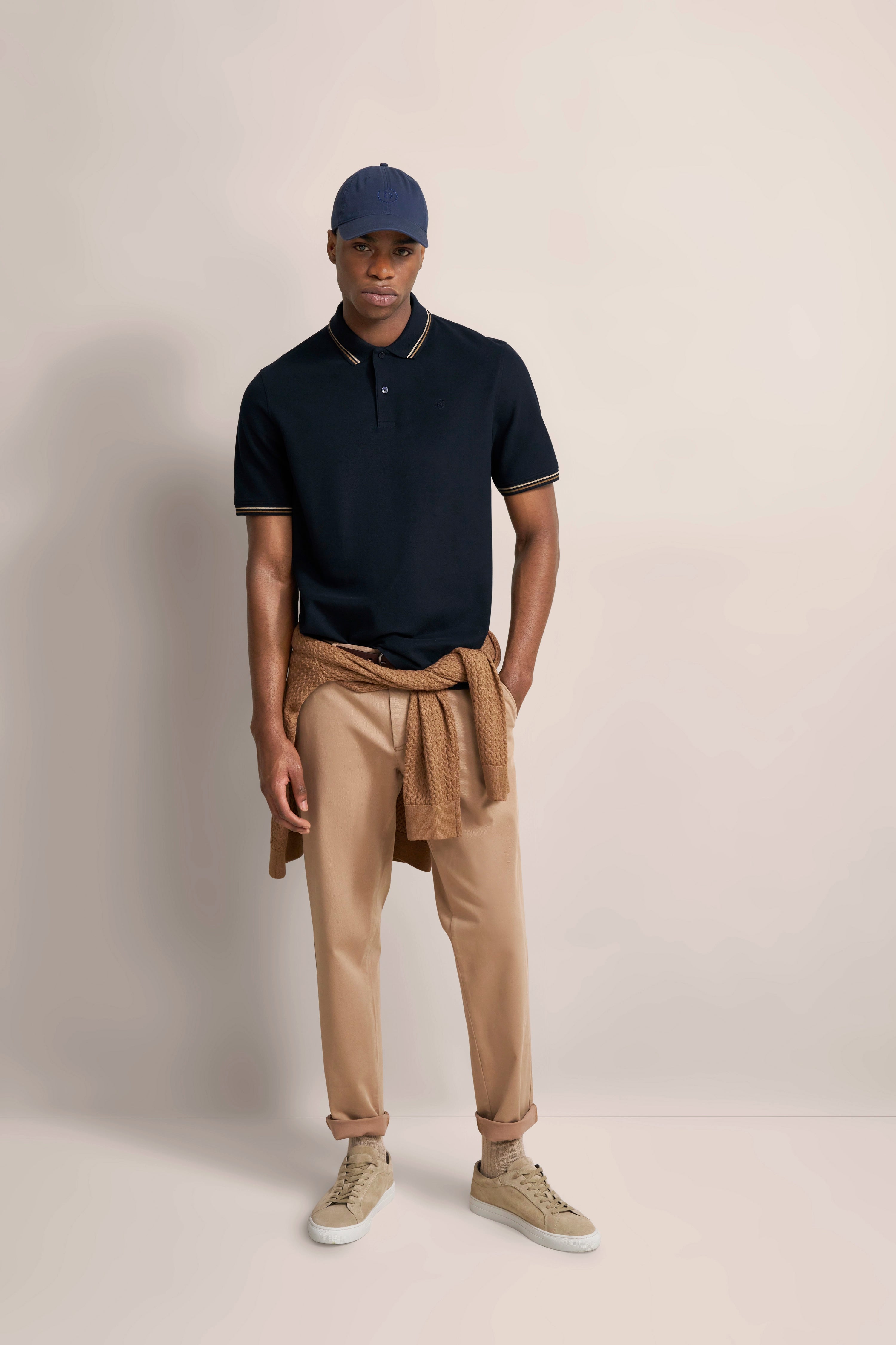 Model in navy Polo, beige Chinos, Strickjacke um die Hüfte, Basecap. Lässiges Steh-Posing.