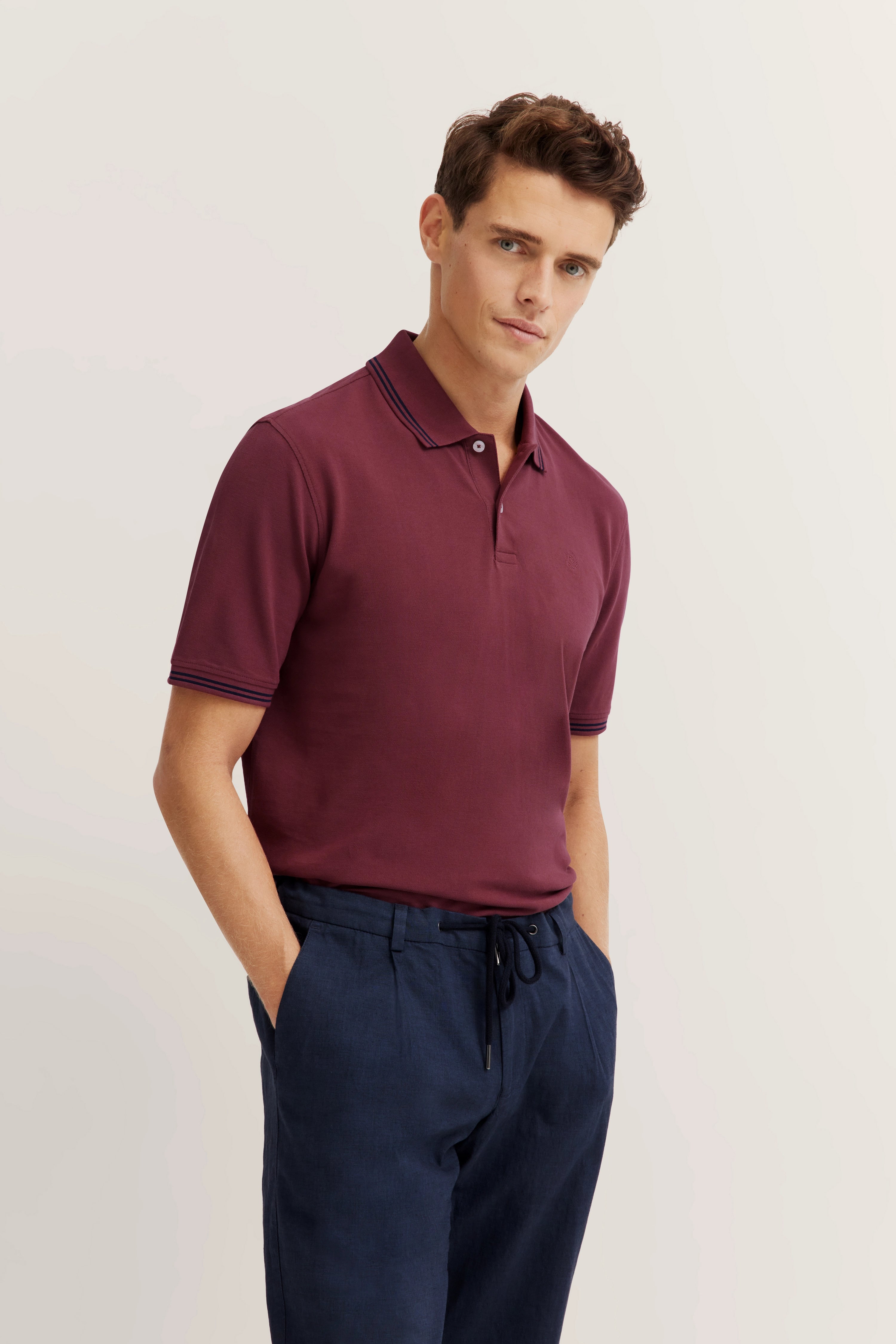 Model in burgundy Polo-Shirt, navy trousers, casual pose, Hände in den Taschen.