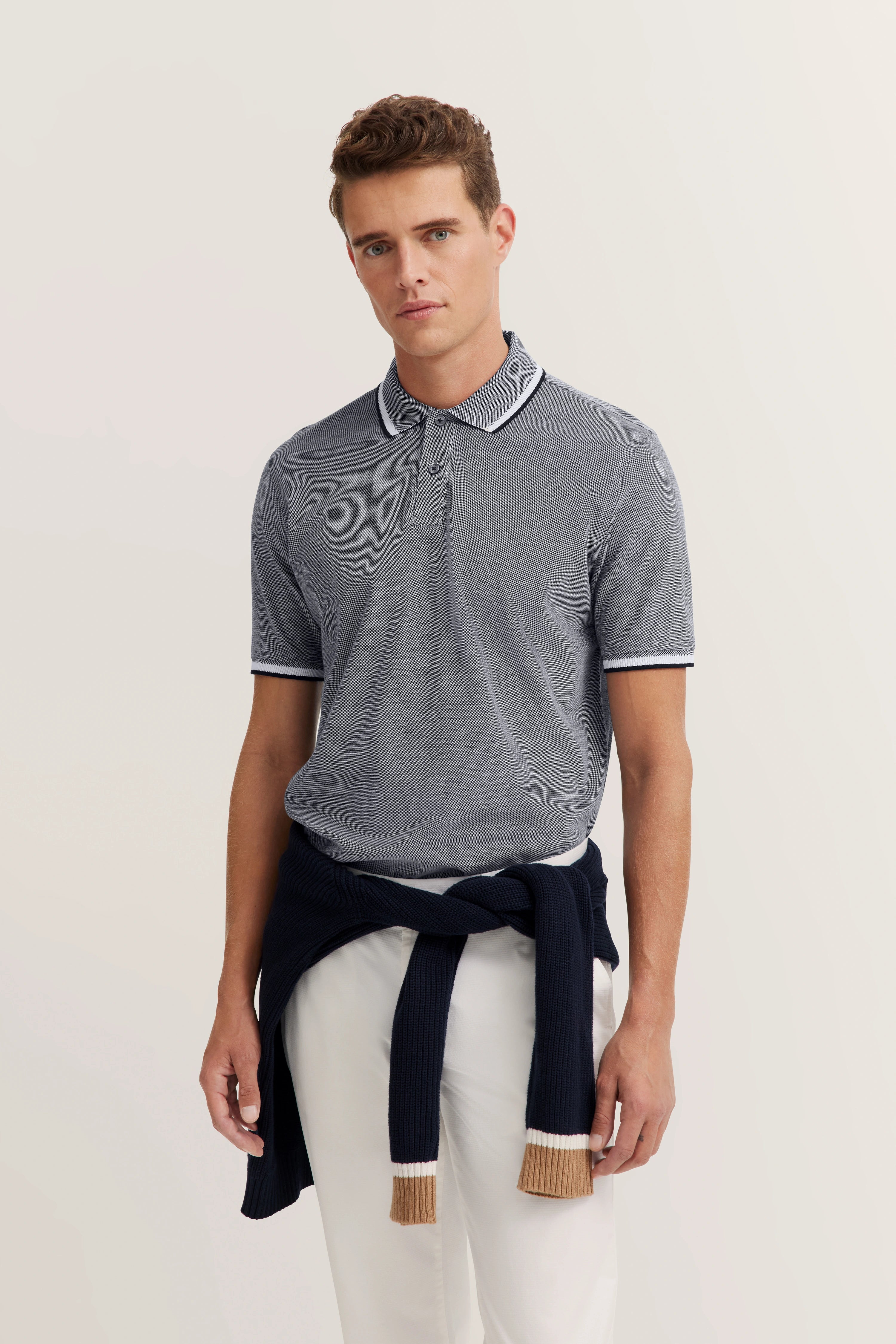 Gray polo shirt with white trim, navy sweater around waist, white background. Model stands relaxed.