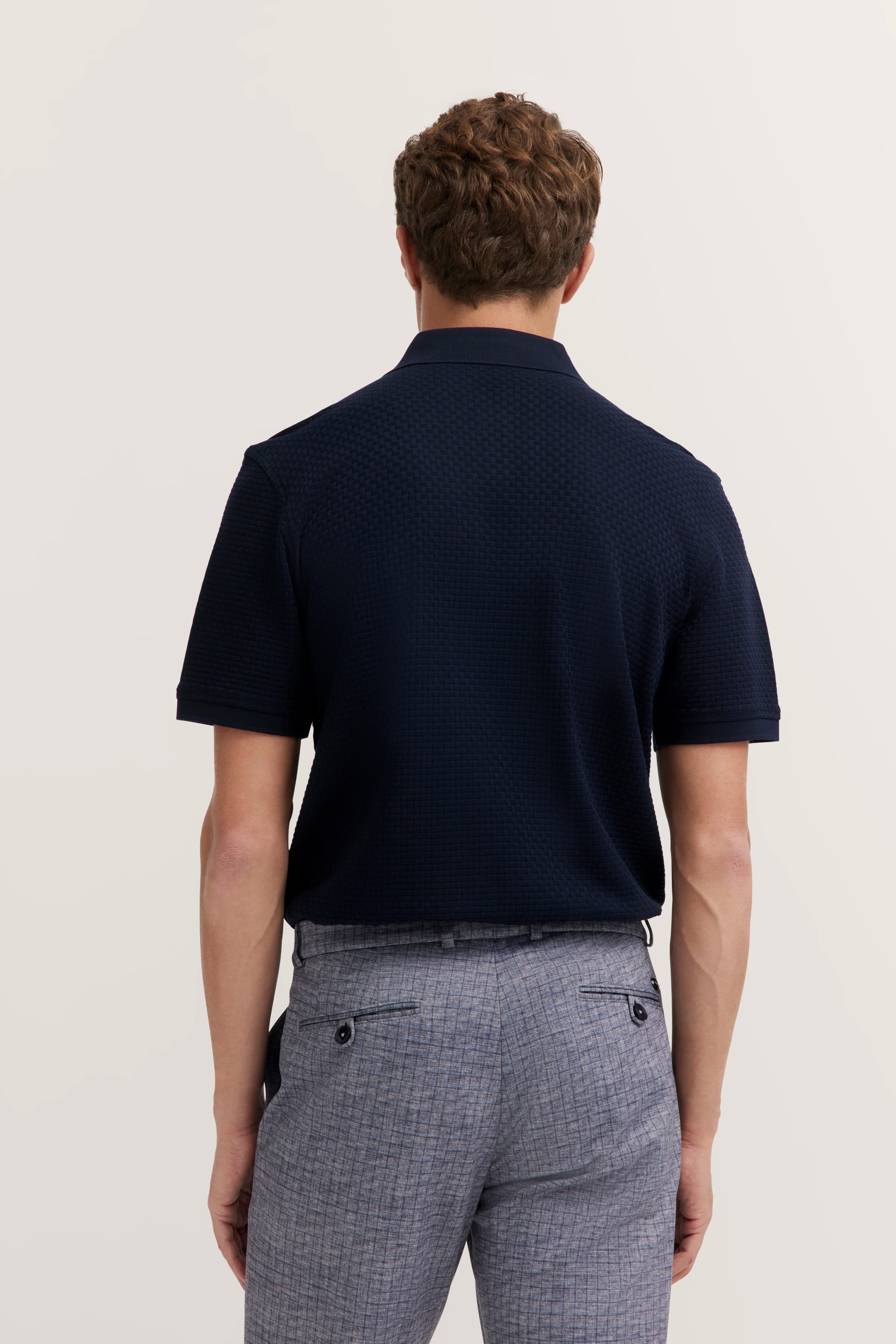 Navy textured polo, grey checkered pants, model back view, relaxed pose.