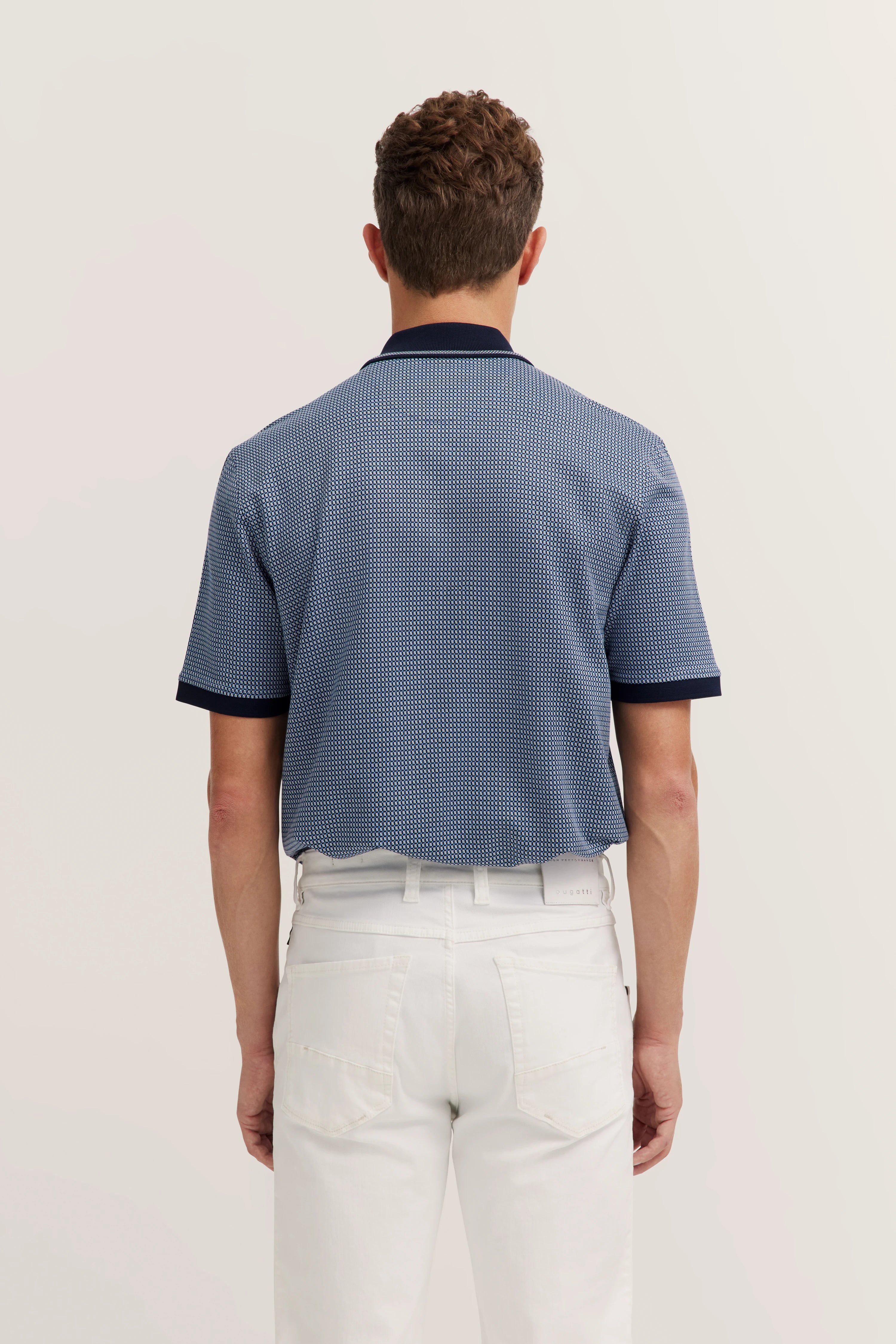 Rear view model in blue patterned bugatti polo, white pants, standing casually.