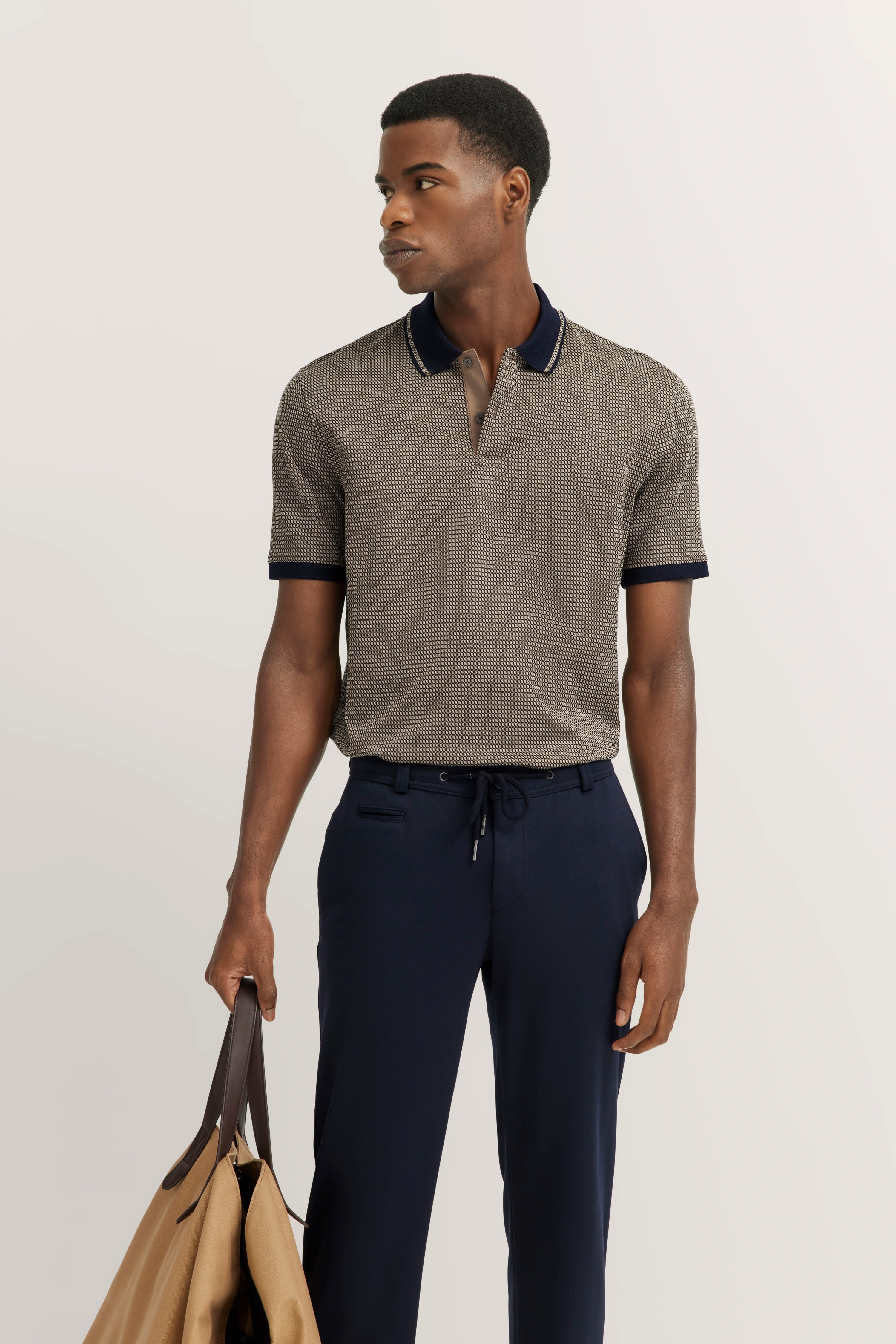 Model in patterned bugatti polo and navy pants, holding bag. Relaxed pose, looking sideways.