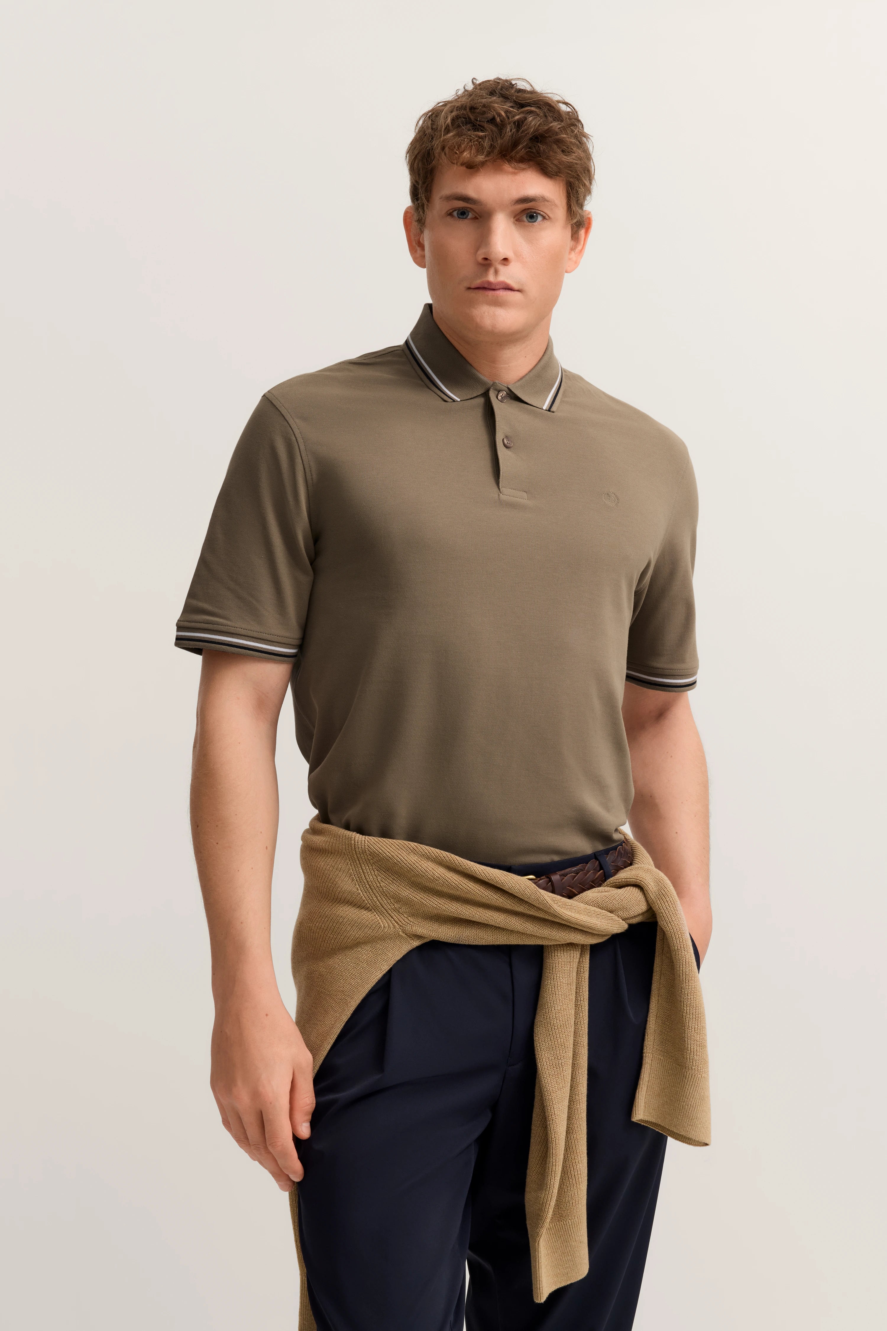 Model in olivgrünem Polo und blauer Hose, beige Pullover um die Taille gebunden.