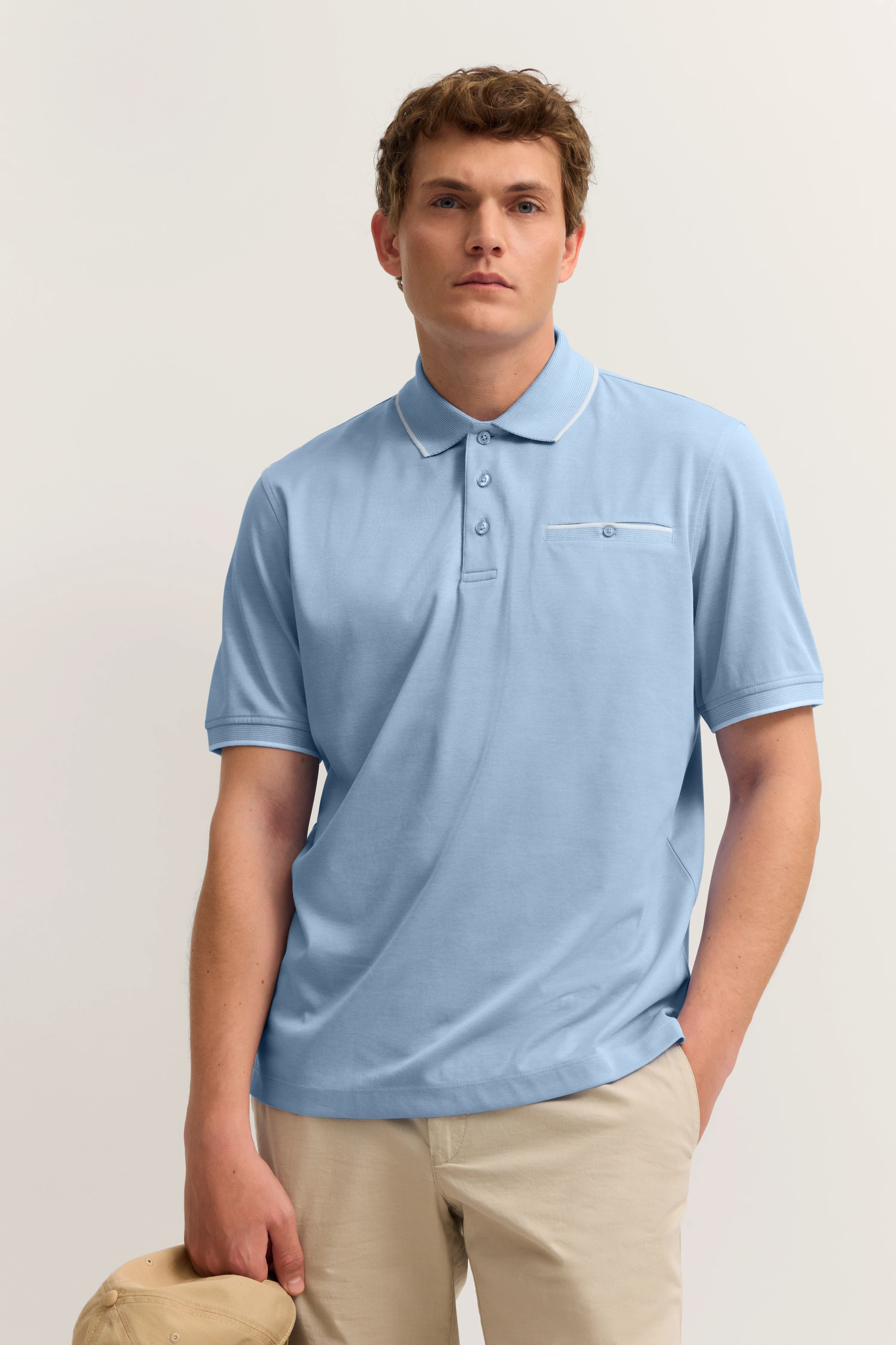Model in hellblauem bugatti Polo und beigen Hosen, legeres Posing, entspannt mit Mütze in der Hand.