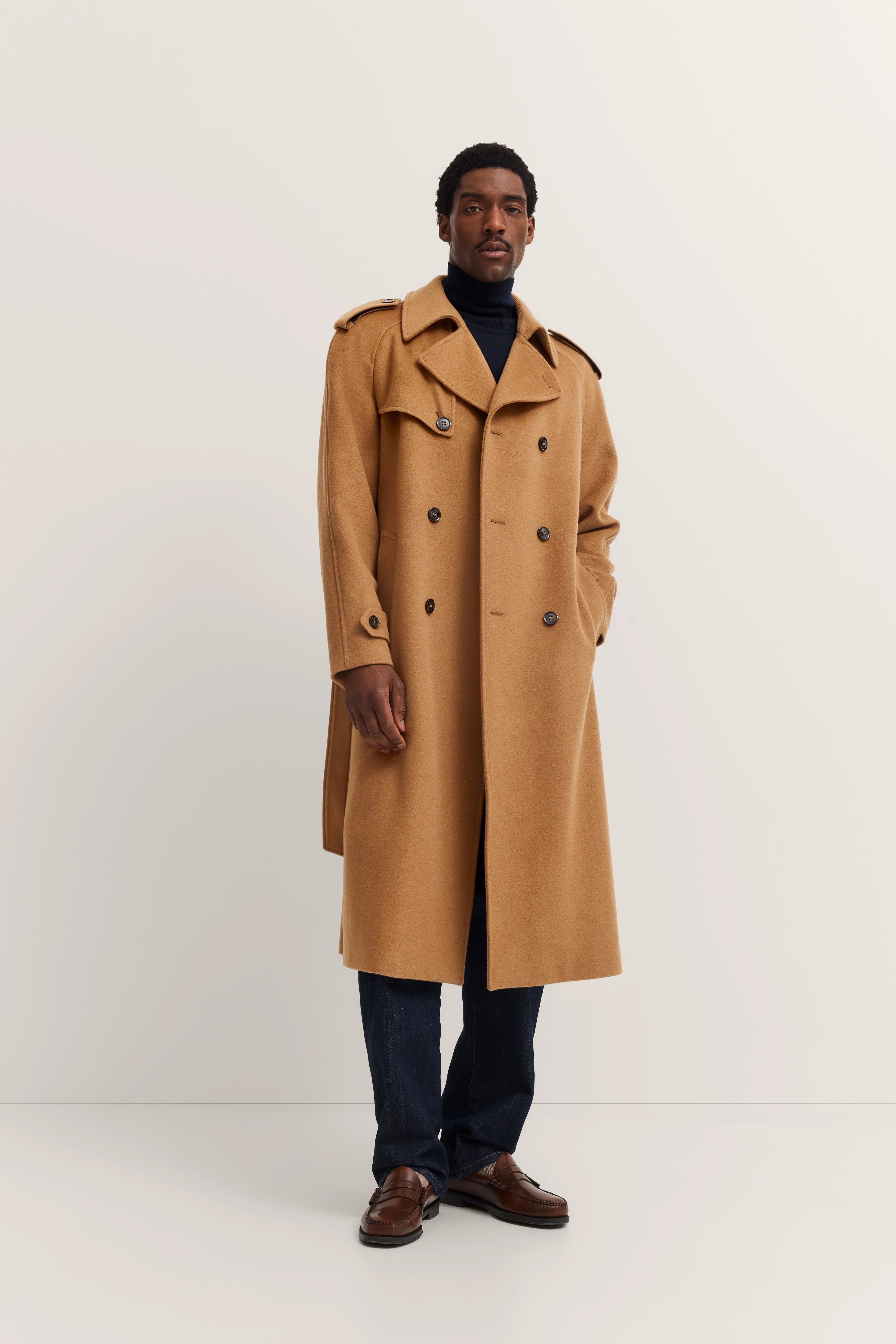 Model in camel trench coat, navy turtleneck, dark jeans, brown leather shoes, exuding relaxed sophistication.