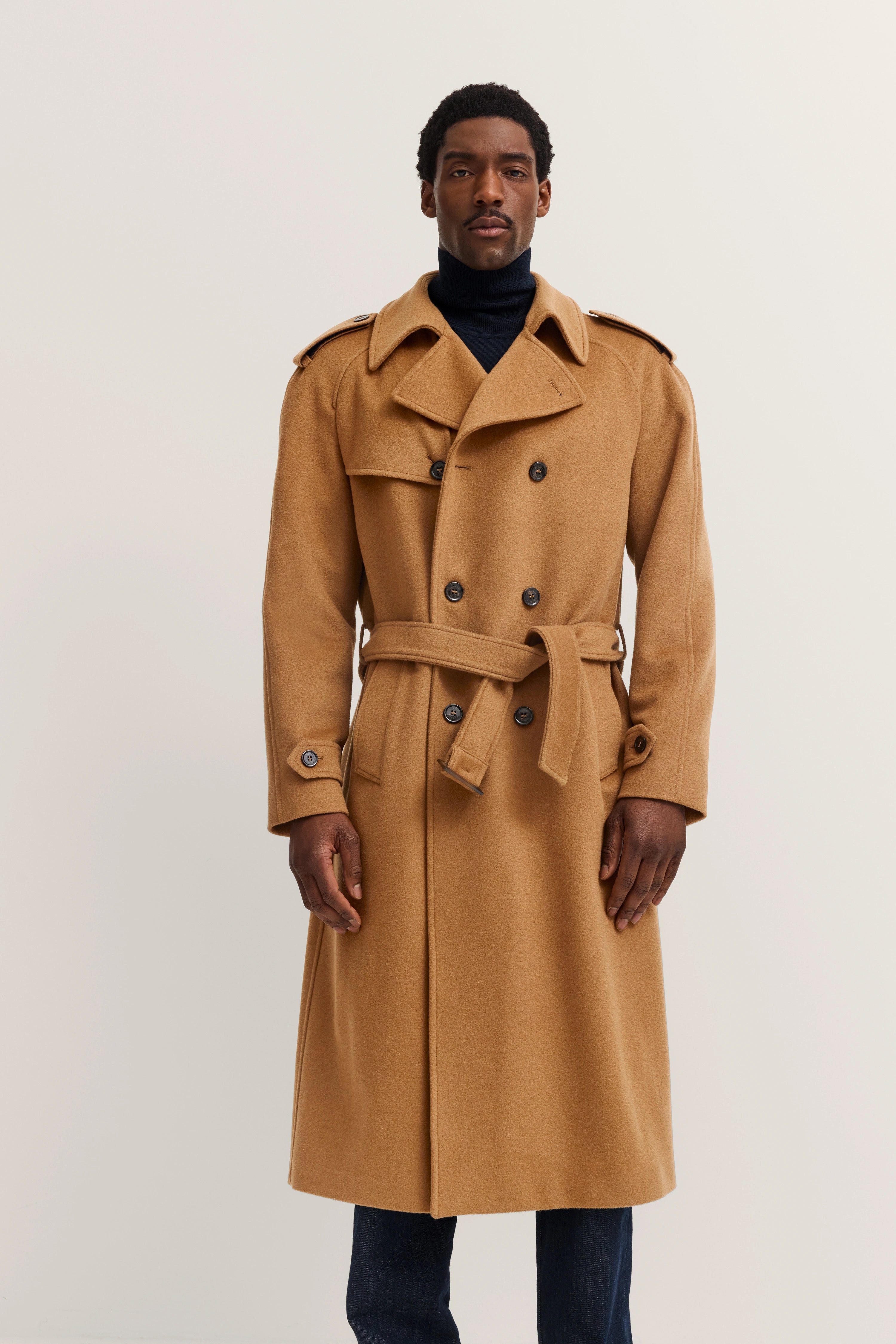 Model in camel bugatti trench coat, belted, black turtleneck, dark blue jeans, exuding timeless style and elegance.