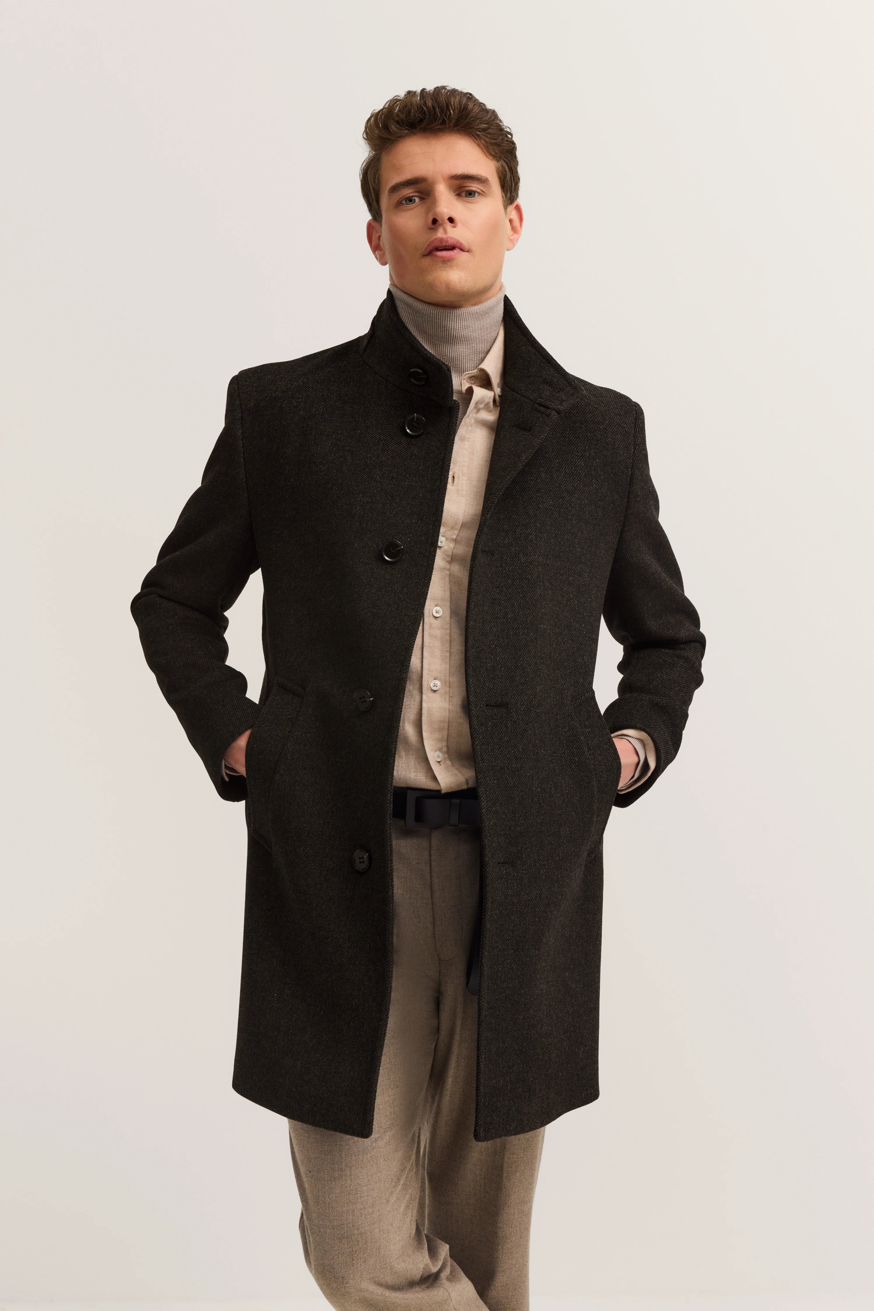 Model in bugatti-jas, donkergrijs, met beige overhemd en broek, nonchalante pose, handen in zakken.