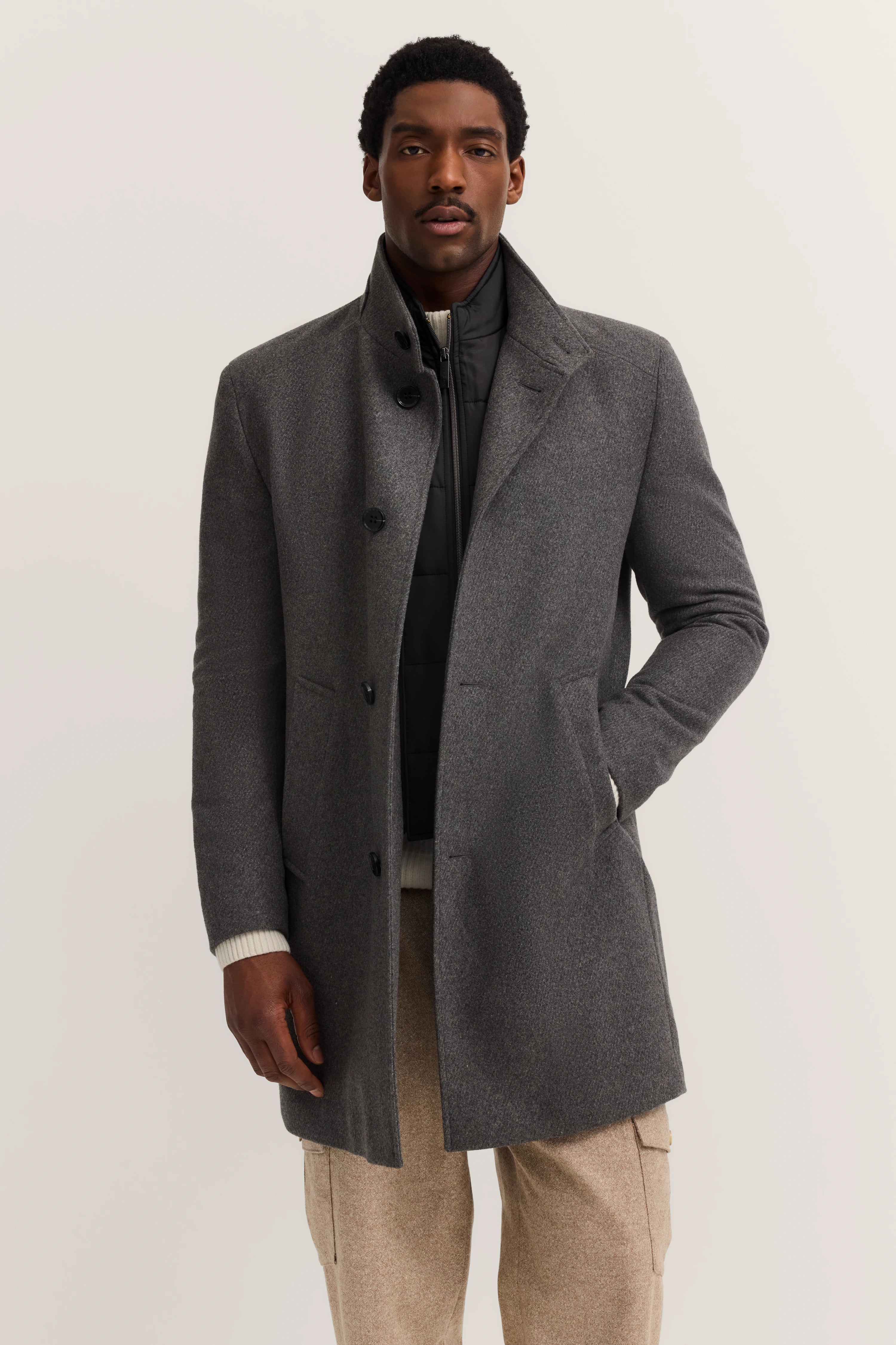 Grauer bugatti Mantel, schwarzer Steppjacke, beige Hose, Modelbild mit entspannter Haltung.