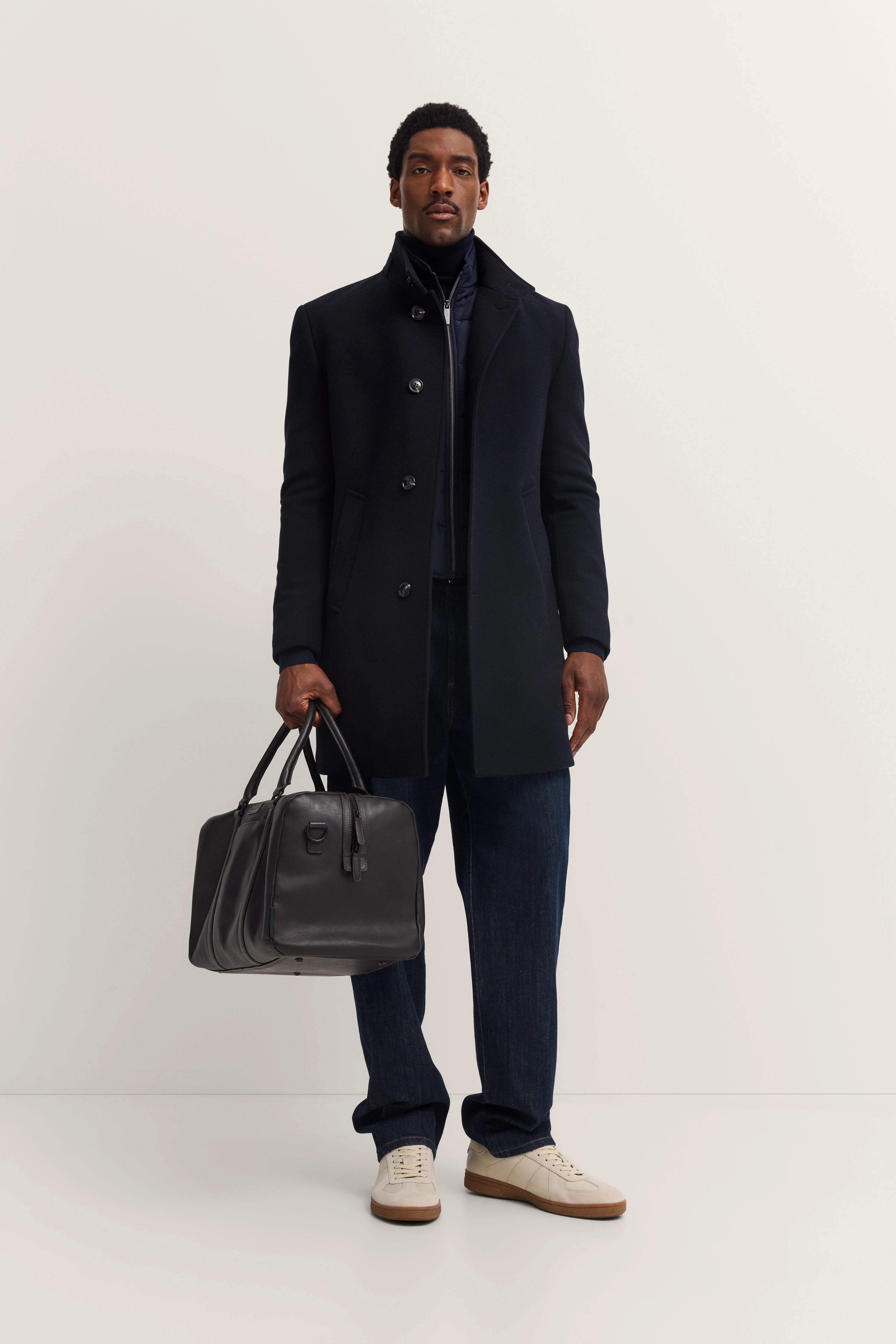 Model trägt schwarzen bugatti Mantel, blaue Jeans, weiße Sneaker und schwarze Tasche, frontal stehend.