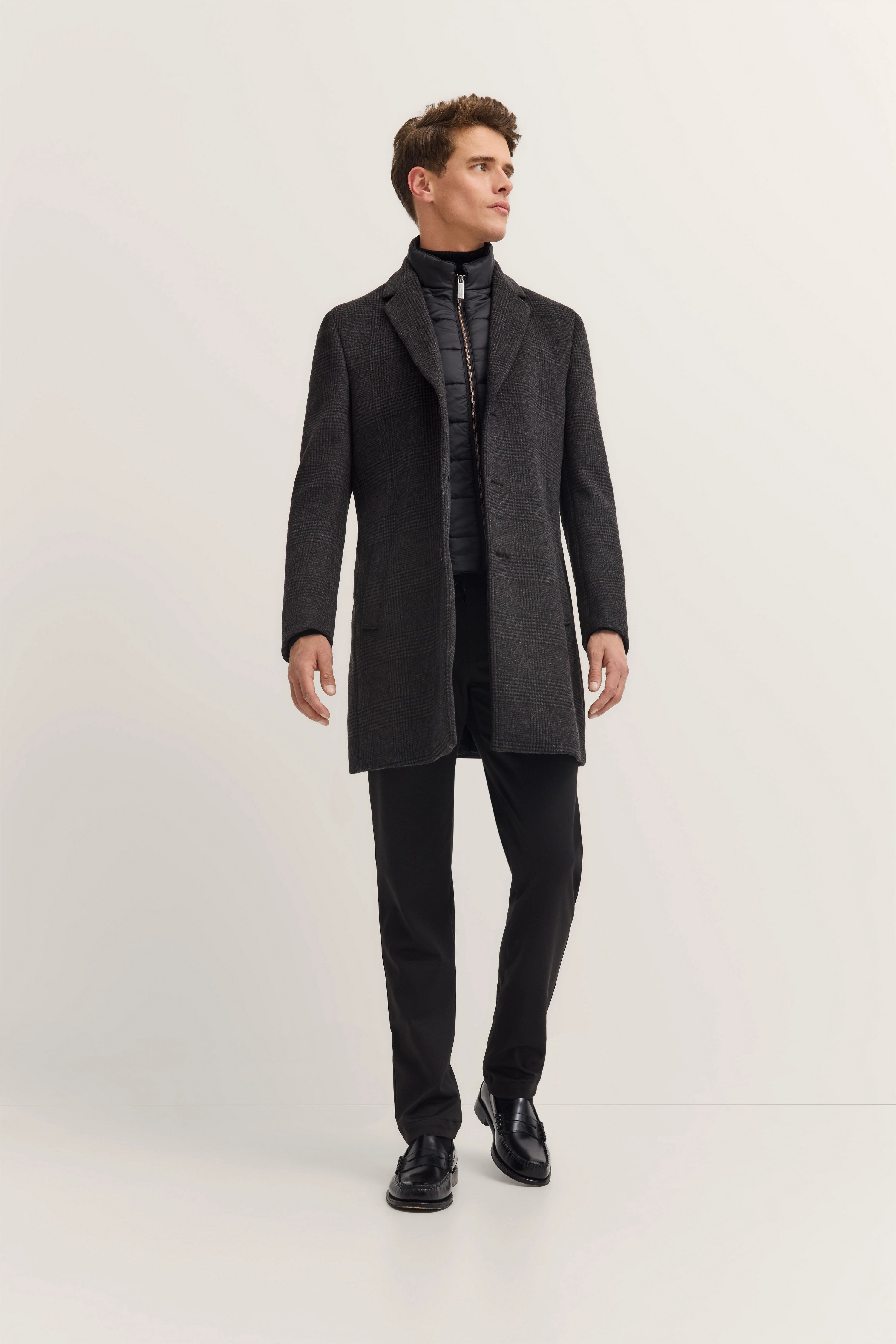 Model in elegantem bugatti Outfit mit grauem Mantel, schwarzem Steppjacke, schwarzer Hose, stehende Pose.