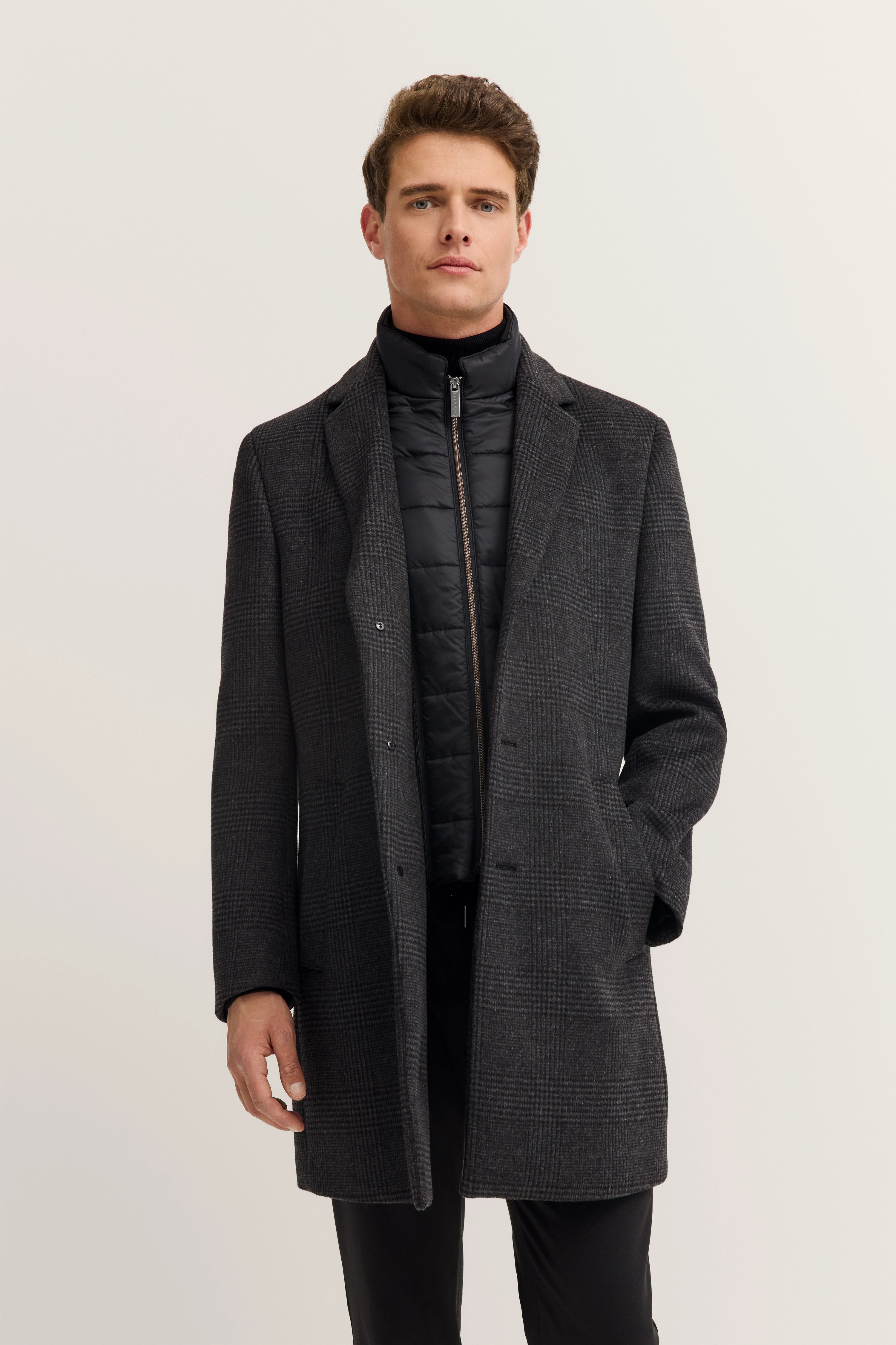 Model trägt grauen bugatti Mantel mit Karomuster über schwarzer Steppjacke, frontal stehend.