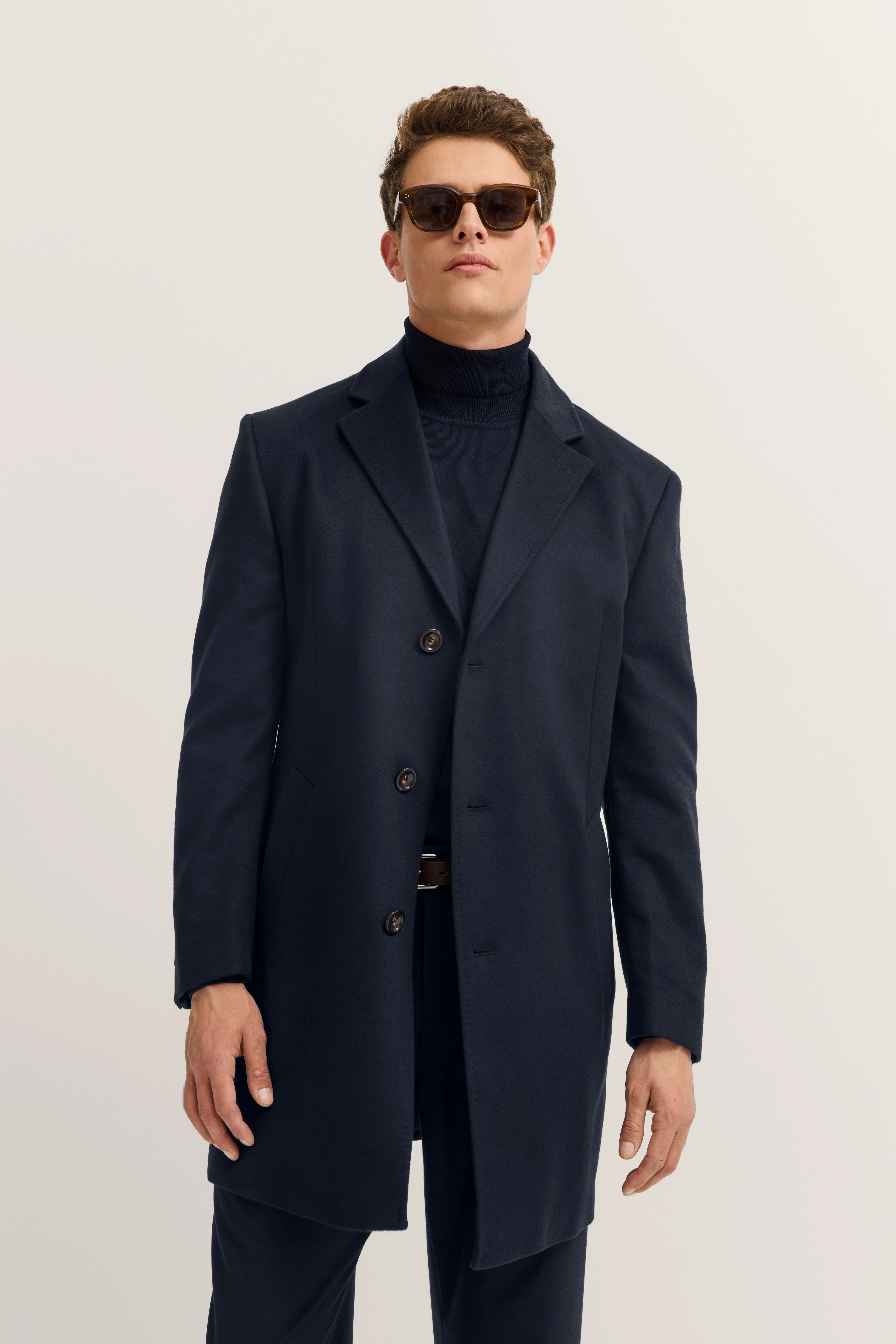 bugatti Herrenoutfit: dunkelblauer Mantel, Rollkragen, Model mit Sonnenbrille, entspannter Stand.