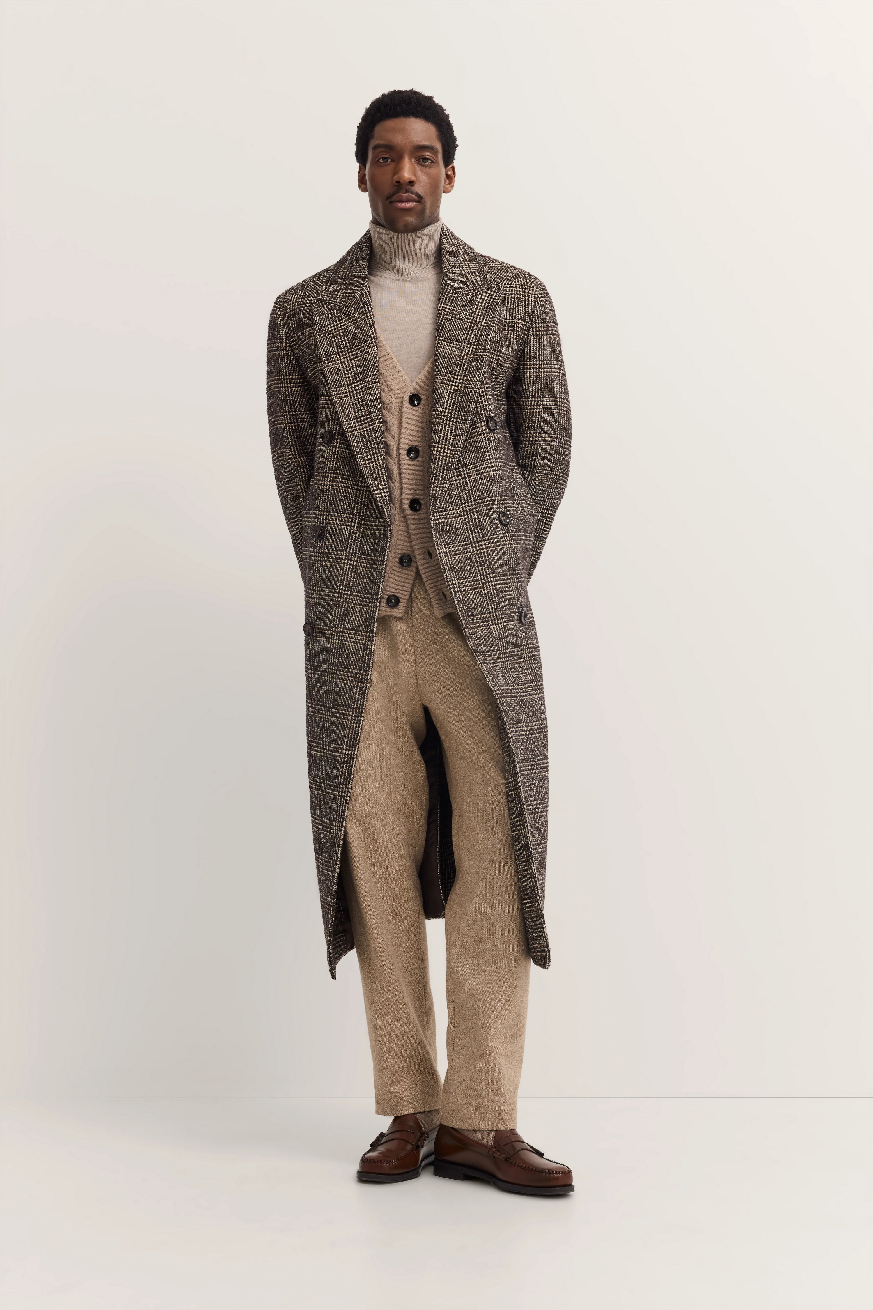 Elegantes Model in kariertem bugatti Mantel, beiger Strickjacke, Rollkragen und Hose, posiert stilvoll.